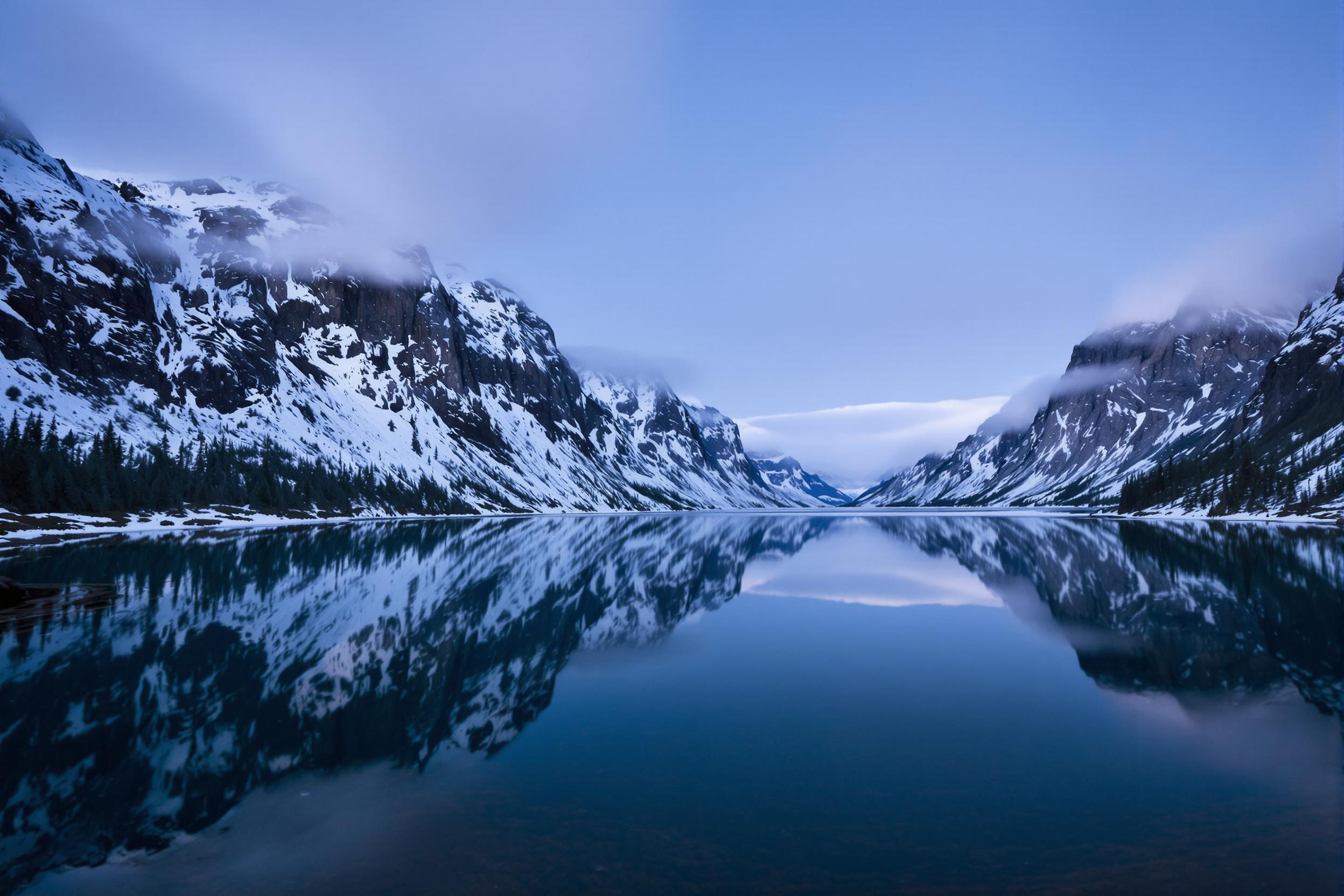 Before sunrise, a tranquil fjord perfectly mirrors its steep granite cliffs under the dim blue-hour sky. Gradient hues fade from deep navy to powder blue across the serene water. Snow-dusted cliffs pierce cloud layers while scattered evergreen trees accentuate the rugged terrain. The still water offers a pristine reflective symmetry, epitomizing natural serenity.