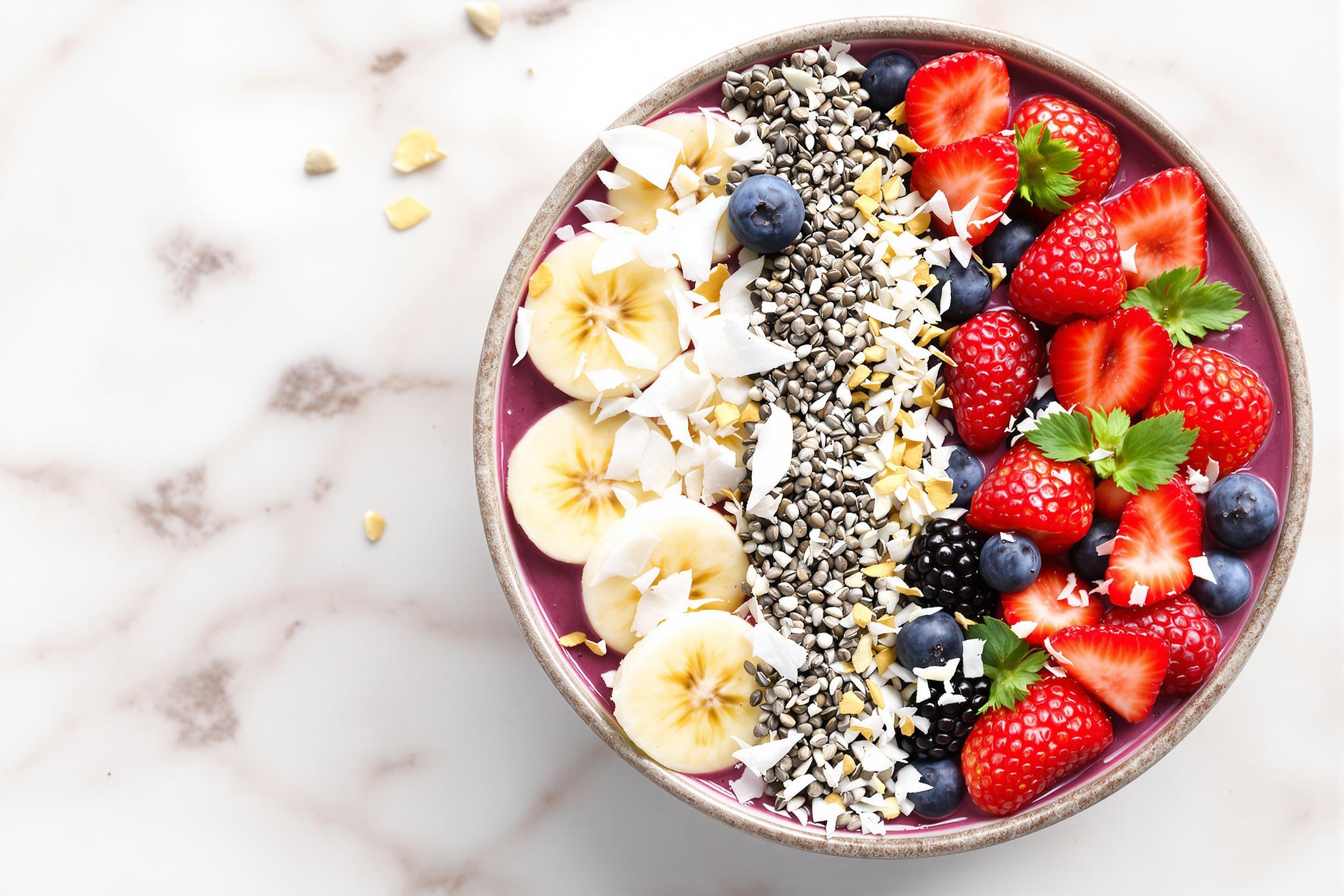 Colorful and nutritious smoothie bowl topped with an array of fresh berries, sliced banana, chia seeds, and coconut flakes. Captured from above on a light marble surface, showcasing the vivid colors and textures of this healthy breakfast option.