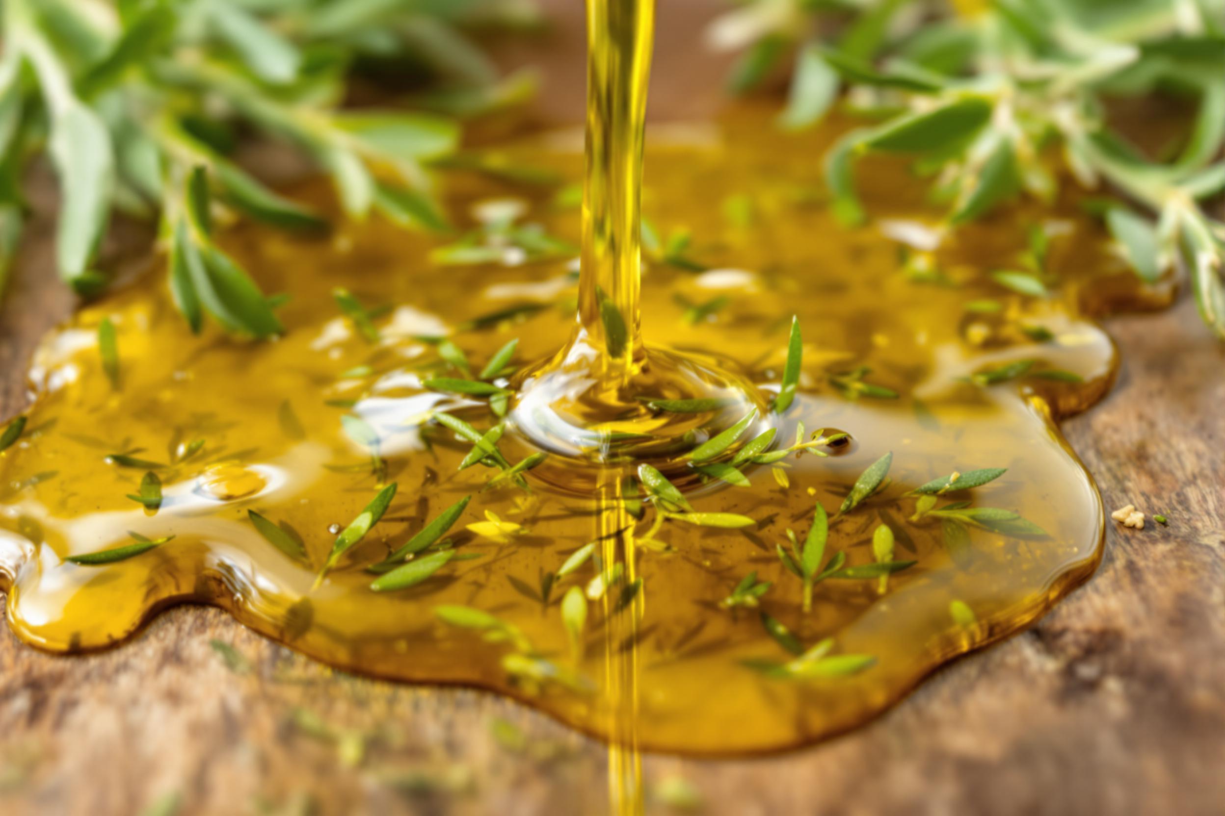 A mesmerizing close-up of golden herb-infused olive oil being drizzled onto a rustic wooden surface. Sprigs of fresh rosemary, thyme, and sage float in the stream, creating an aromatic and visually striking culinary scene.