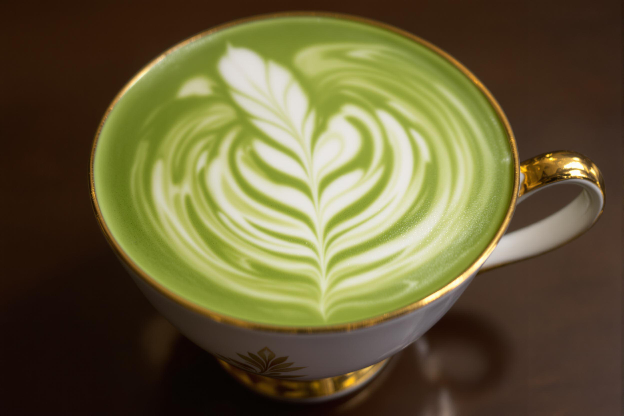 Exquisite matcha latte art showcased in a delicate vintage porcelain teacup. Swirls of vibrant green matcha contrast beautifully with creamy white foam, creating an intricate leaf design. Soft, diffused lighting accentuates the cup's ornate gold rim and handle.