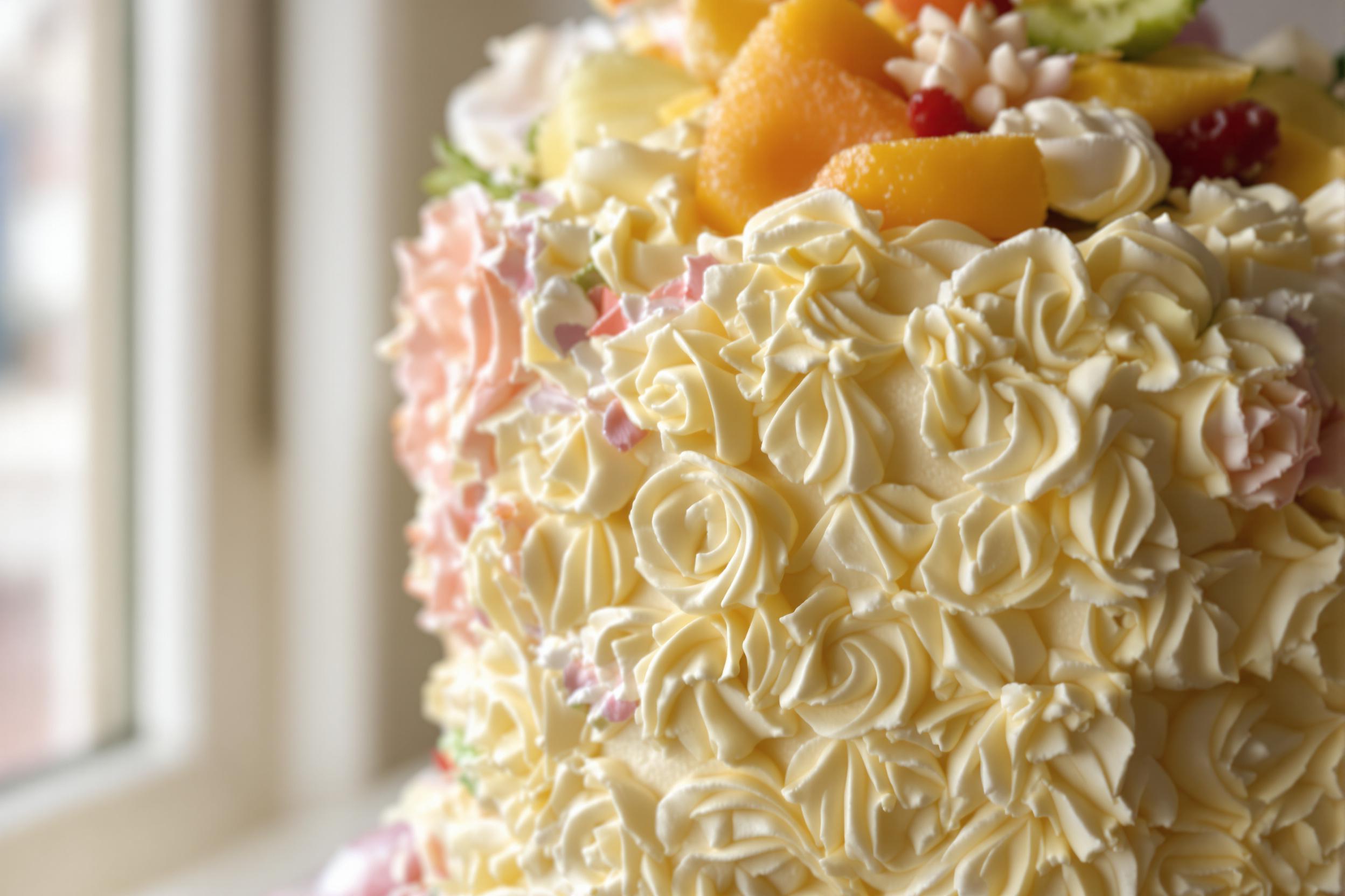 A mouthwatering close-up features a beautifully decorated vanilla cake with rich buttercream frosting. Intricate floral designs in pastel colors cascade down the sides, while fresh fruit sits atop the cake as vibrant accents. The natural light from a nearby window enhances the textures of the frosting, creating a delightful and inviting scene.