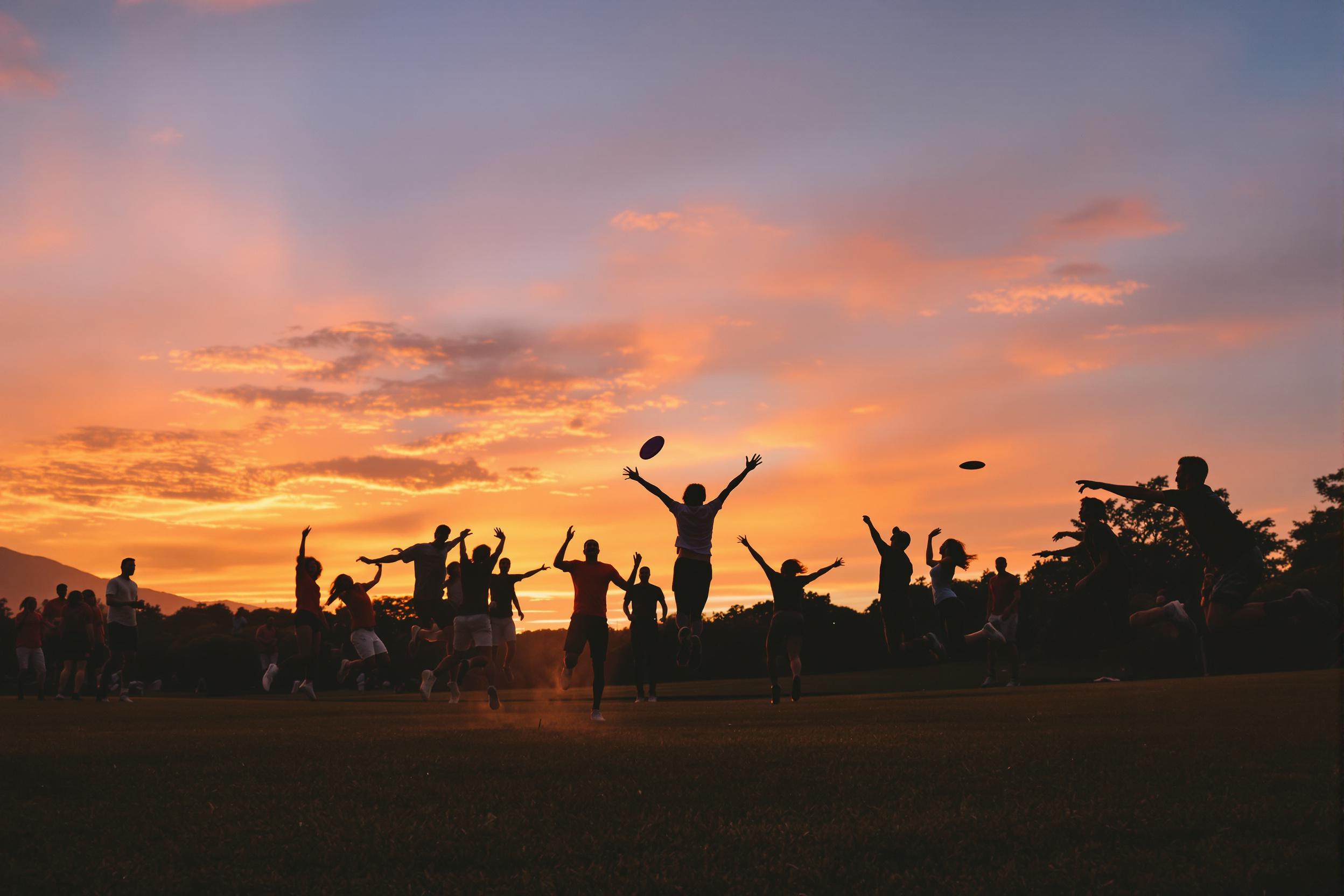 A lively gathering unfolds as a group of friends enjoys an energetic game of frisbee in a sprawling park during sunset. The sky is painted in shades of orange and pink, casting a warm glow on the grassy field. Laughter fills the air as they dive and leap, creating dynamic silhouettes against the vibrant backdrop. This scene captures a moment of carefree fun and camaraderie.