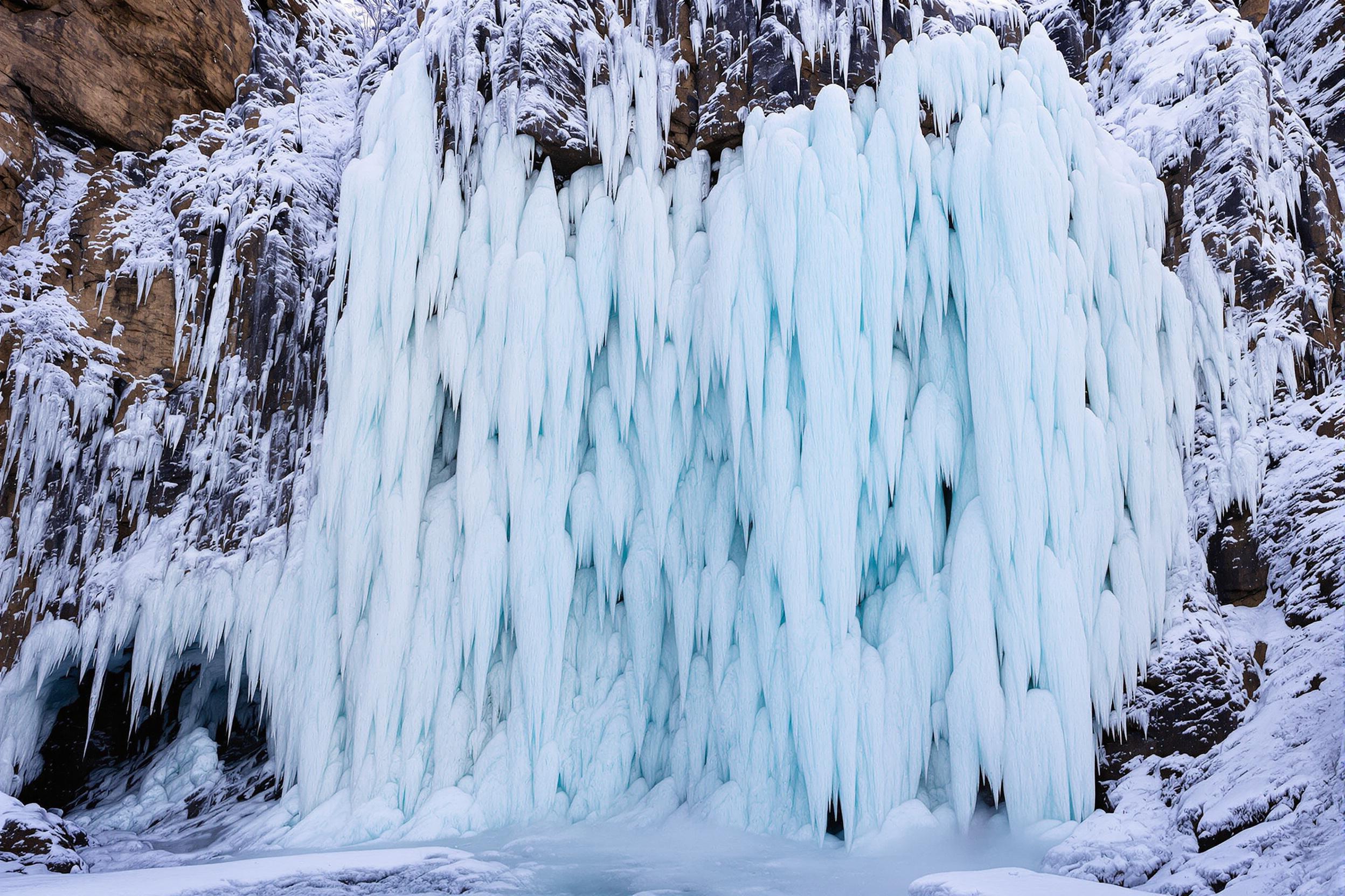 Towering frozen waterfalls cascade down jagged cliffs, forming intricate ice sculptures beneath pale winter sunlight. Delicate icicles glisten in cold air, accentuating complex patterns of ice layers. Frosty mist drifts gently from faintly visible cavernous spaces below. Snow covers surrounding rocks, creating a stark contrast to the blue-tinted crystalline formations.