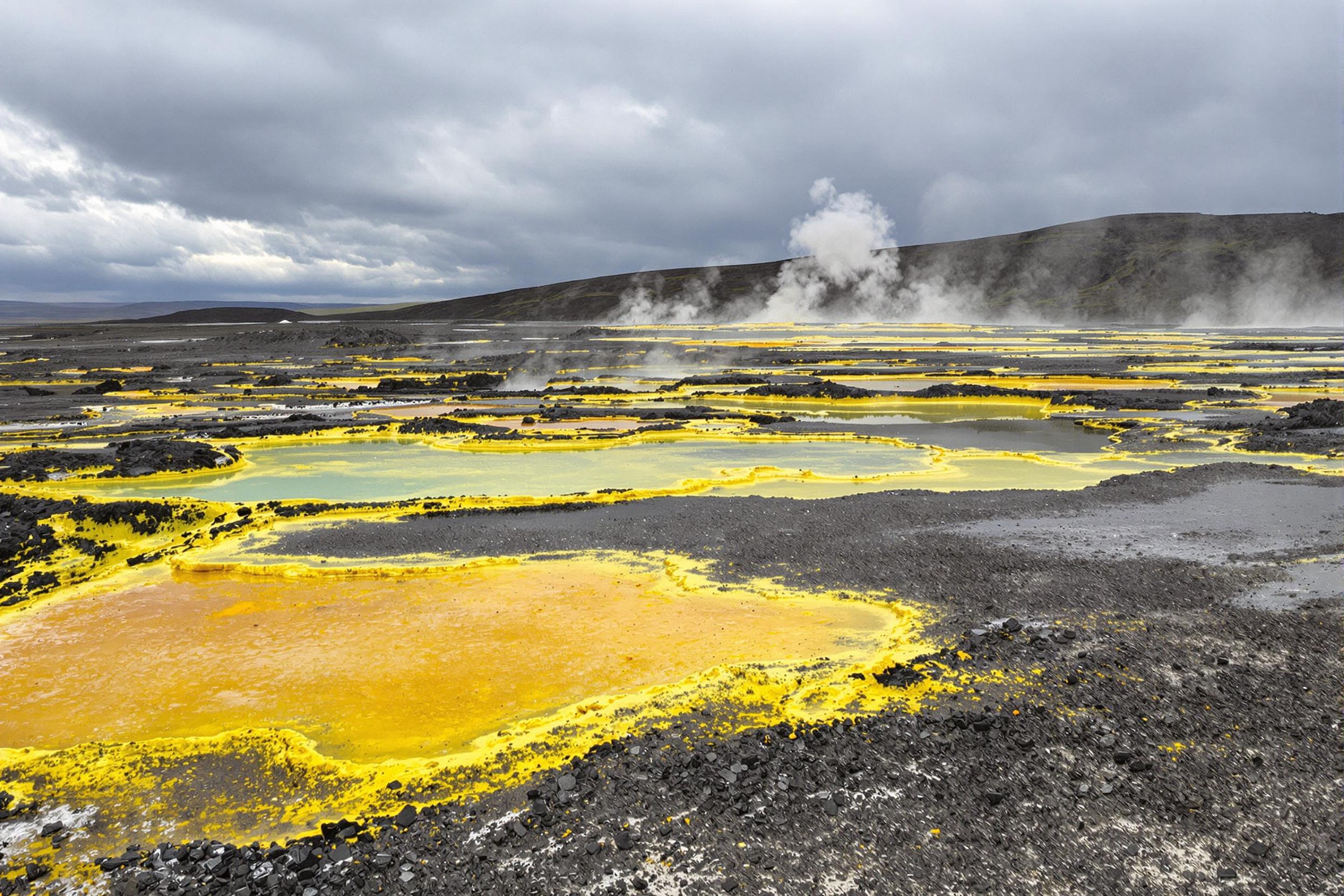 Sulfuric steam vents rise against a desolate, rocky geothermal landscape under turbulent gray skies. Vibrant yellow sulfur deposits contrast with the ashy terrain, creating sharp boundaries of color and texture. Pools of iridescent, mineral-rich water reflect scattered light, while wisps of steam distort the stark horizon. The atmospheric setting highlights Earth's raw, primal energy.