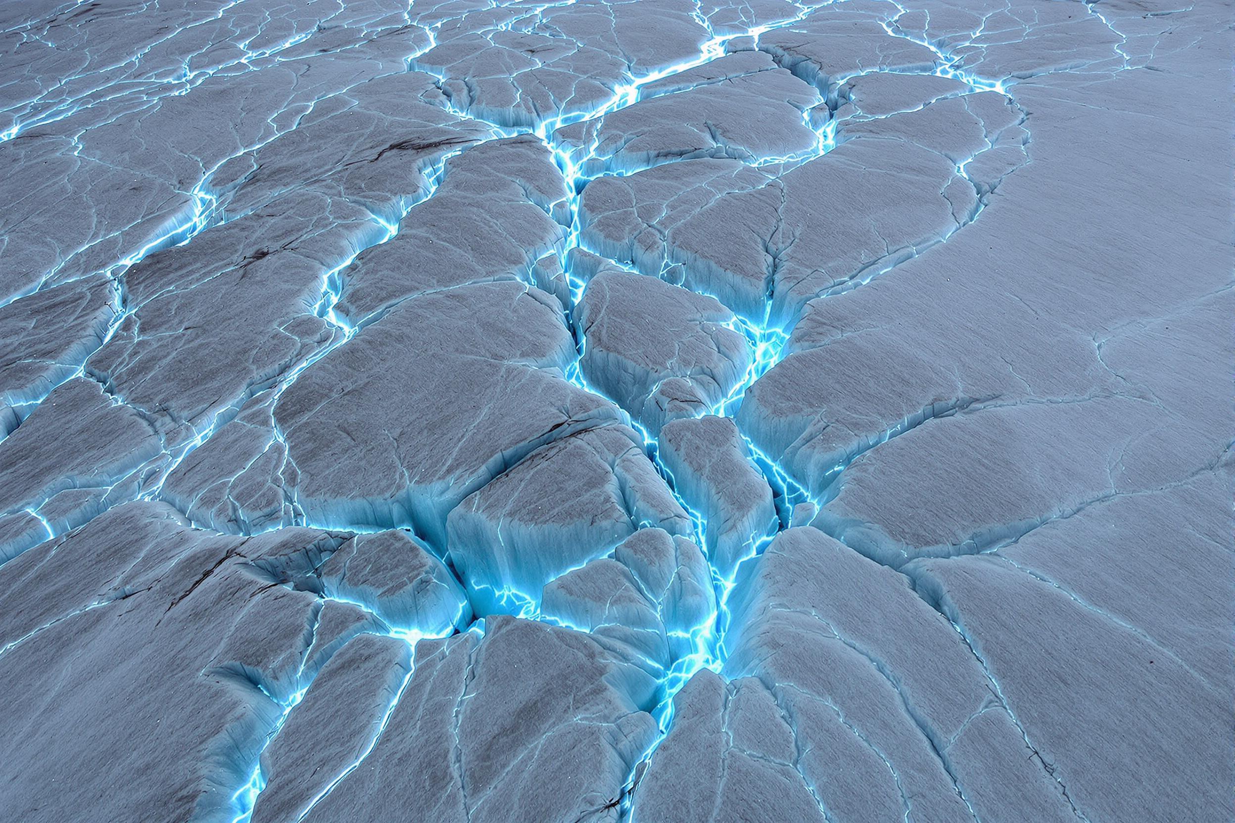 An expansive aerial view reveals a glacier's jagged surface etched with deep crevices and winding veins of brilliant blue ice. The cracked expanse captures texture and depth, with glowing white ice contrasting against frigid shadowy recesses. Subtle mist enhances the icy atmosphere under overcast light, amplifying the scene's dramatic scale.