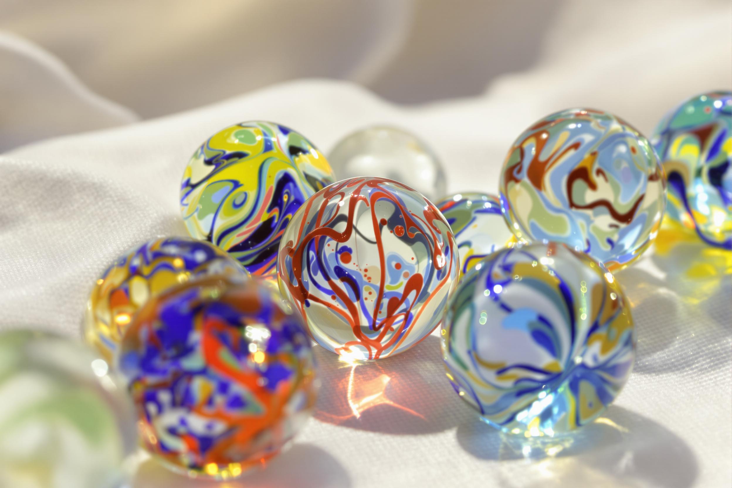 A close-up display of intricate glass marbles resting on a soft, textured fabric background. The marbles showcase a spectrum of vivid colors, including deep blues, bright reds, and sunny yellows, enhanced by the gentle midday light. Each marble's surface gleams, reflecting subtle patterns and swirls unique to its design. The composition invites a closer look at their craftsmanship, creating a visually engaging arrangement.