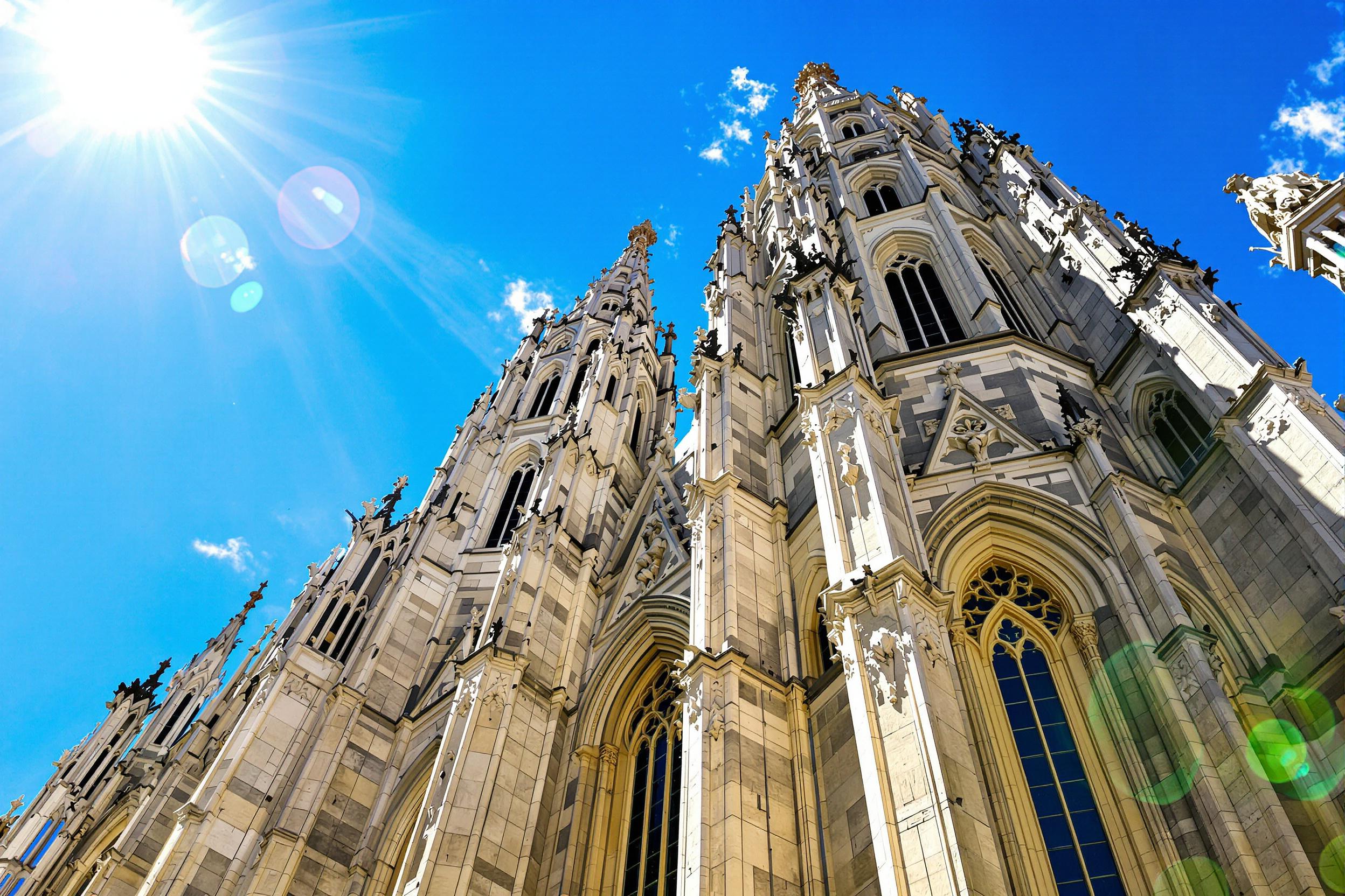 An intricate perspective captures the architectural grandeur of a Gothic cathedral facade under brilliant sunlight. Its towering spires adorned with detailed stone carvings rise against a vivid blue sky. Intricately chiseled gargoyles contrast with the graceful symmetry of stained glass windows peeking through ornate stone frameworks.