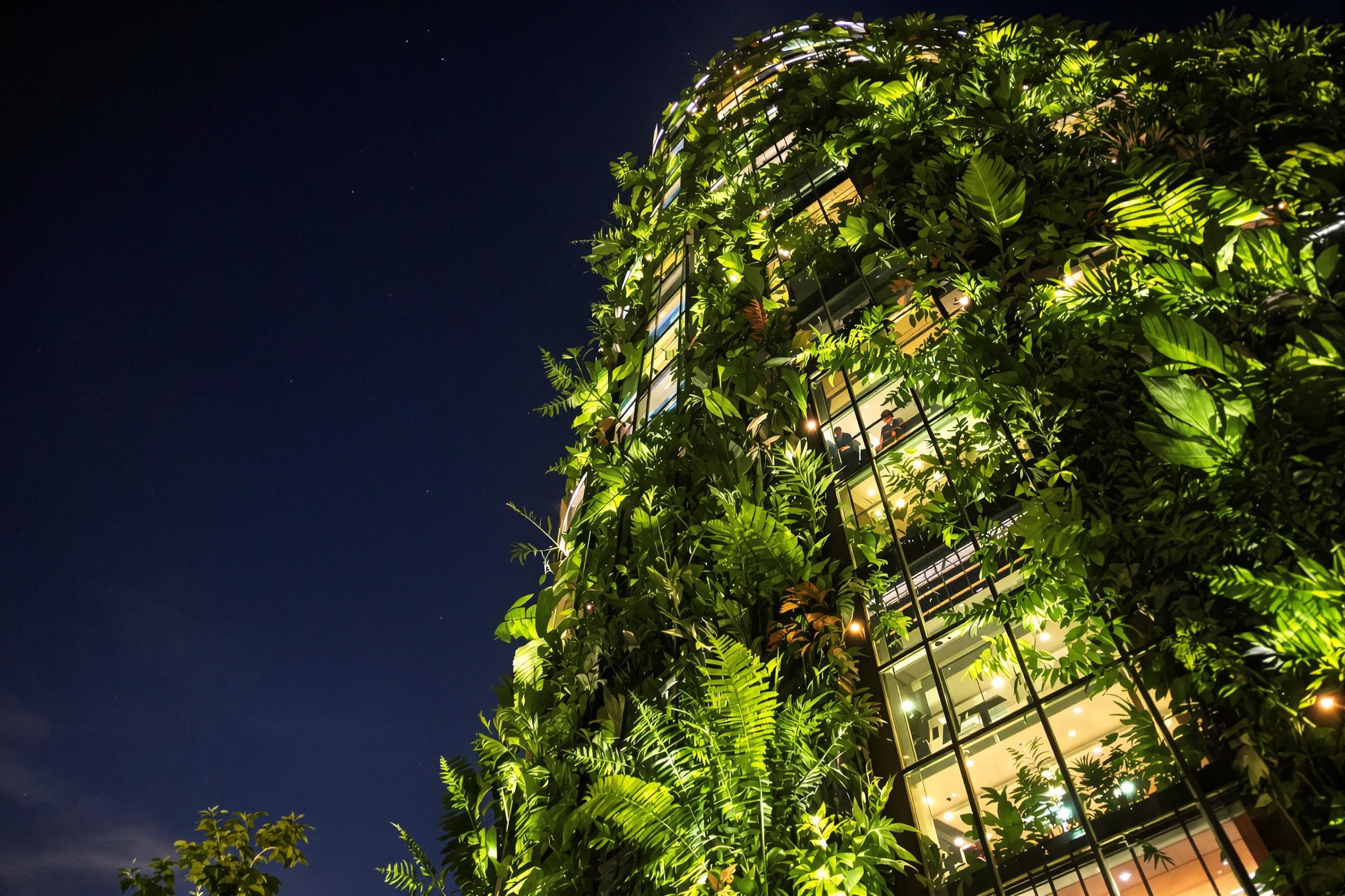 A dramatic worm’s-eye view captures a vertical garden skyscraper against a clear night sky. The building's façade overflows with dense ferns and tropical plants illuminated by warm, soft LEDs. Reflective glass panels contrast the greenery, mirroring distant city lights below. Sharp textures define the structure while the lush vegetation softens it delicately.