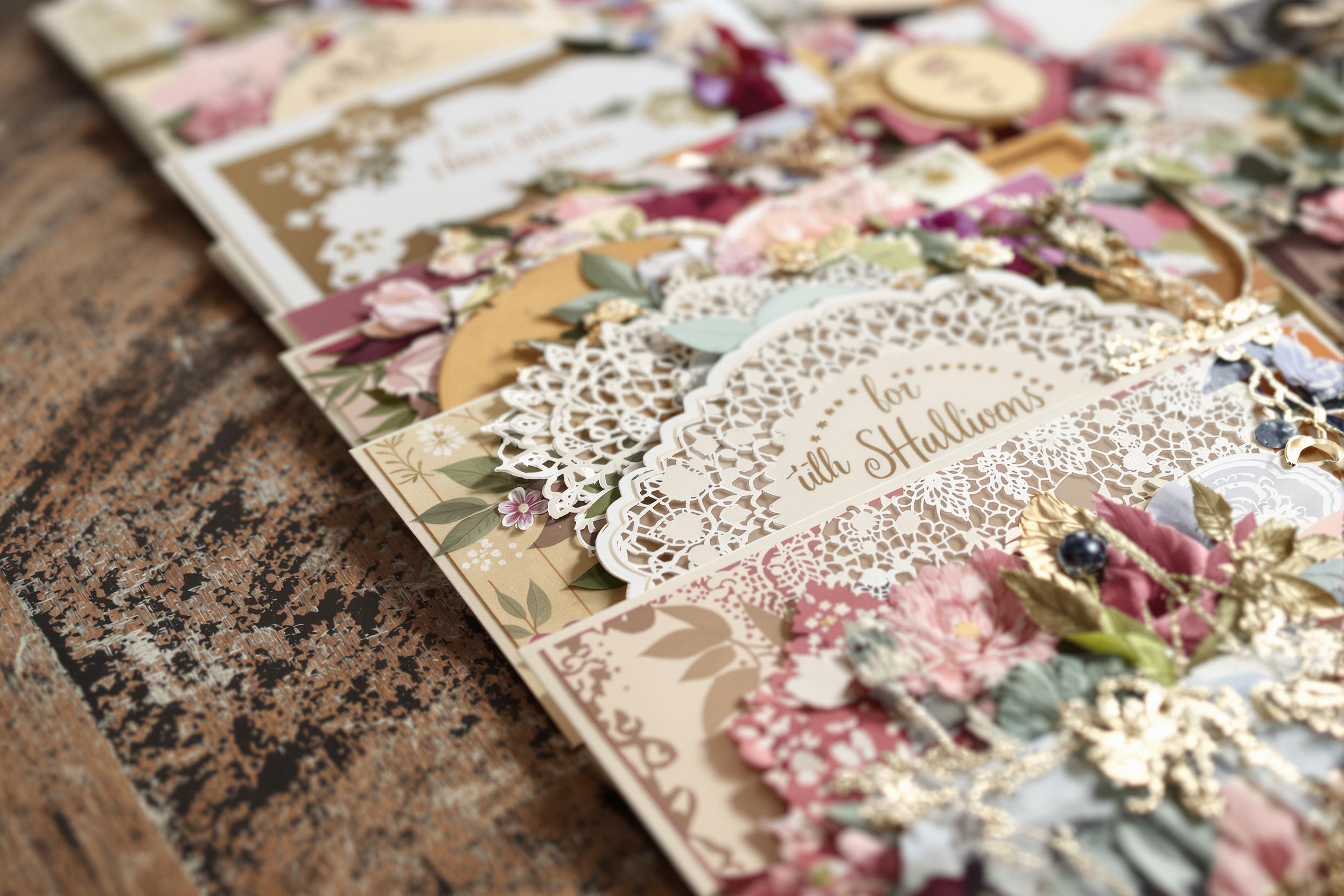 A close-up captures an array of handcrafted greeting cards arranged on a rustic wooden table. Each card features intricate designs, lush colors, and varied textures like lace and floral patterns. Soft, diffused light highlights the glossy finishes and delicate paper, while the blurred background provides depth, focusing attention on the artisanal details that make each card unique.