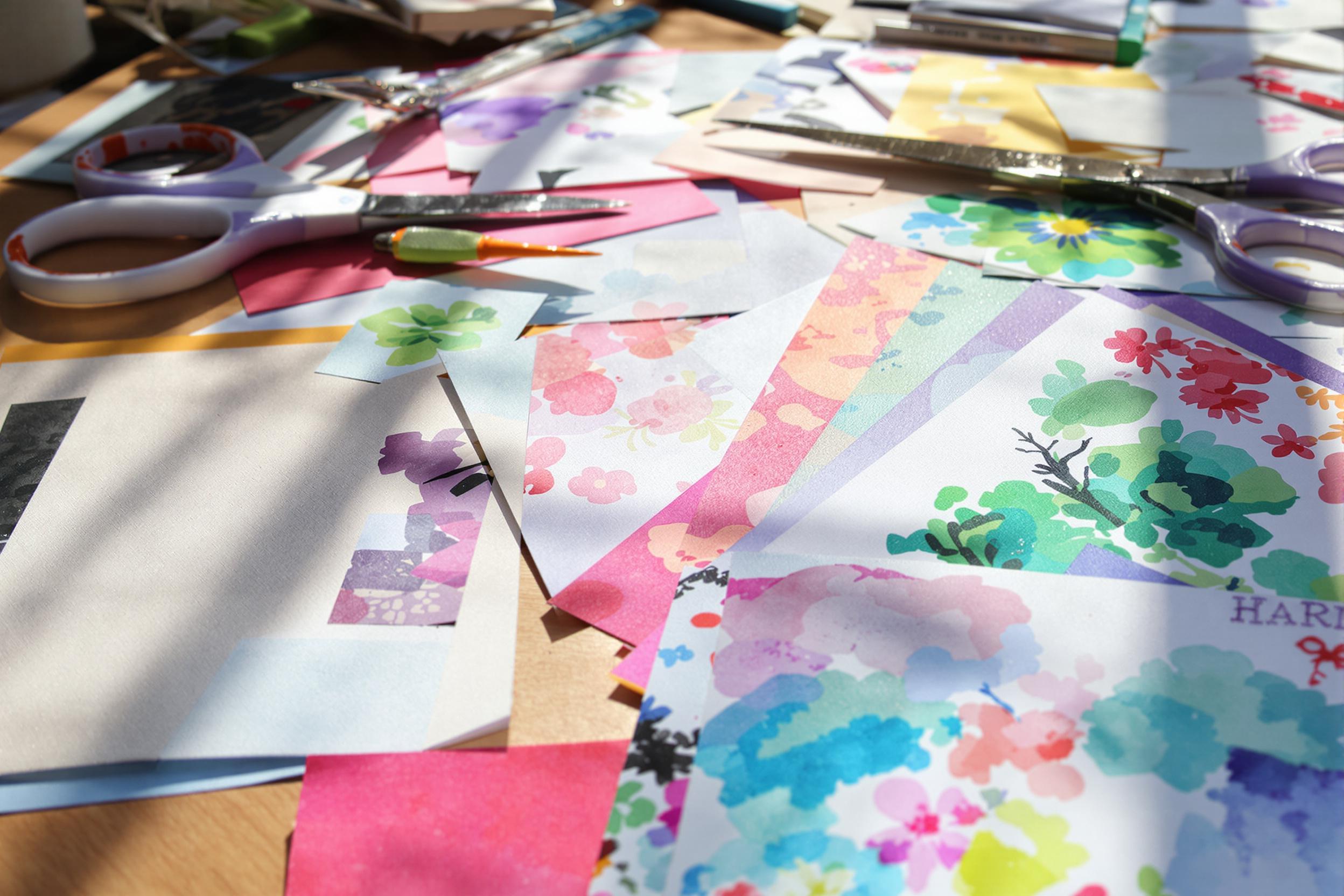 An intimate close-up captures an array of handcrafted papers scattered across a wooden table. Textures range from subtle linen to vibrant watercolor patterns, illuminated by bright midday sunlight. Scissors and other crafting tools are strategically placed, creating a dynamic composition. Soft shadows play across the surface, enhancing the colorful collage of materials.