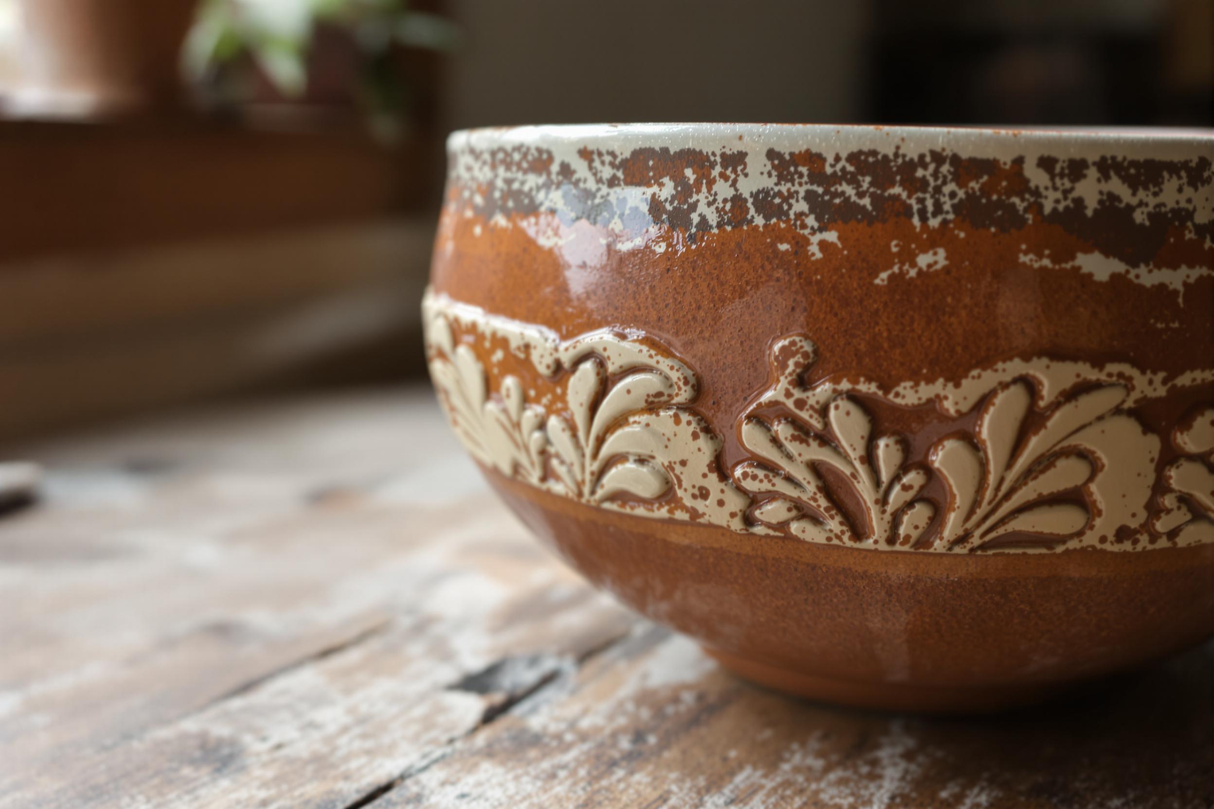 A close-up captures an exquisite handmade pottery bowl resting on a worn wooden table. The bowl's surface displays a rich blend of earthy tones—rustic reds, browns, and softer cream accents—showcasing the artisan's skill. Gentle natural light filters through a nearby window, illuminating the subtle textures and intricate patterns carved into the clay.