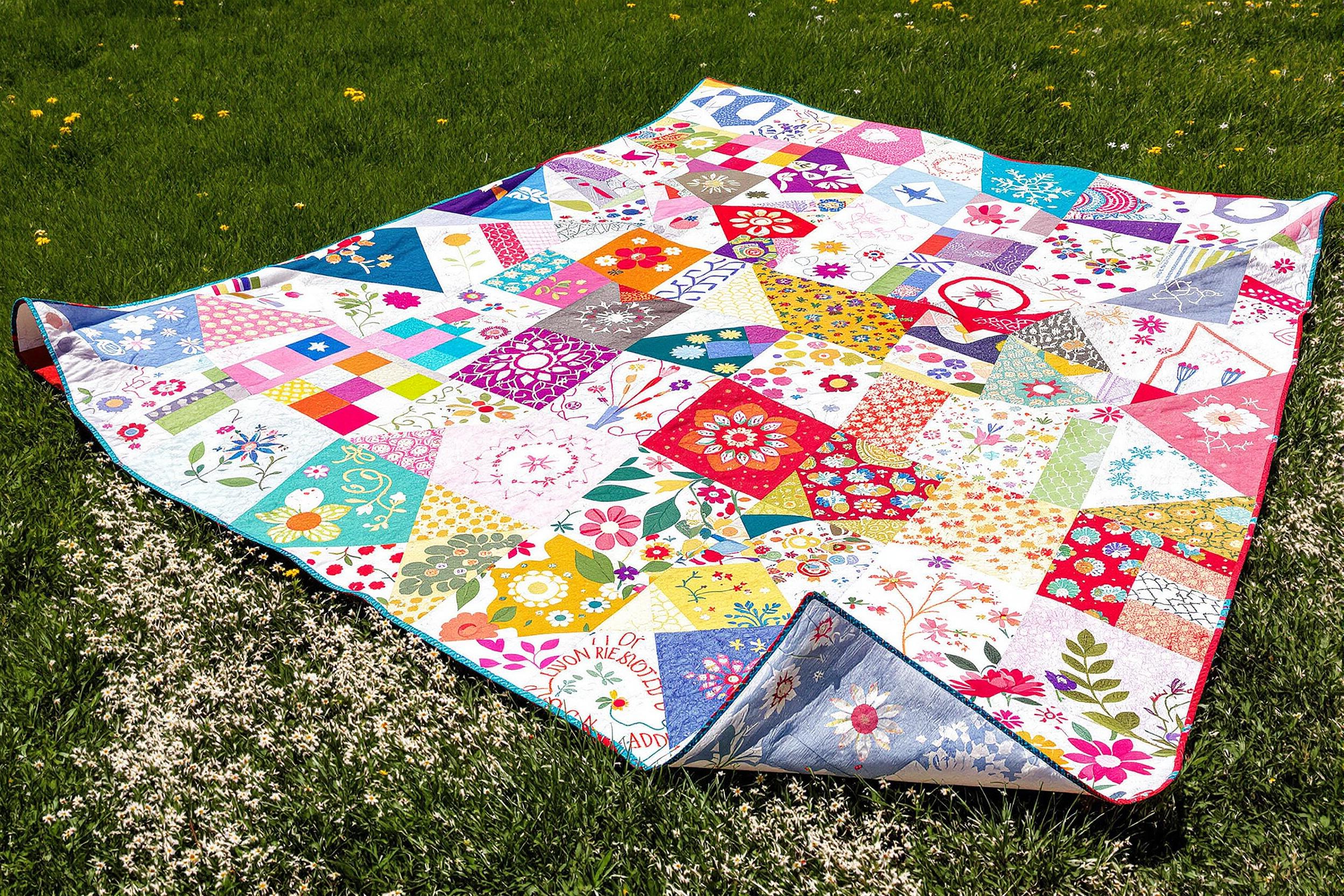 A colorful handmade quilt laid out on a vibrant green lawn presents a patchwork of intricate designs including floral patterns, geometric shapes, and delicate embroideries. Bright sunlight highlights the varied textures while nearby wildflowers add splashes of color. Soft shadows dance across the quilt, enhancing its inviting appearance as a perfect setting for a picnic.