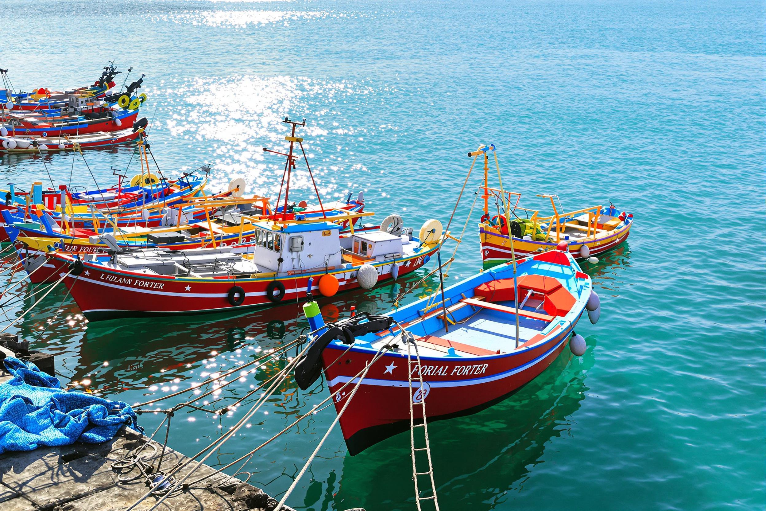 A lively harbor scene showcases an array of colorful fishing boats gently bobbing on the azure waters. The early morning sun casts a soft, warm glow, illuminating the vibrant red, blue, and yellow hulls. Reflections ripple across the water, creating dynamic patterns. Lively ropes and nets are strewn about the docks, enhancing the busy yet peaceful atmosphere.