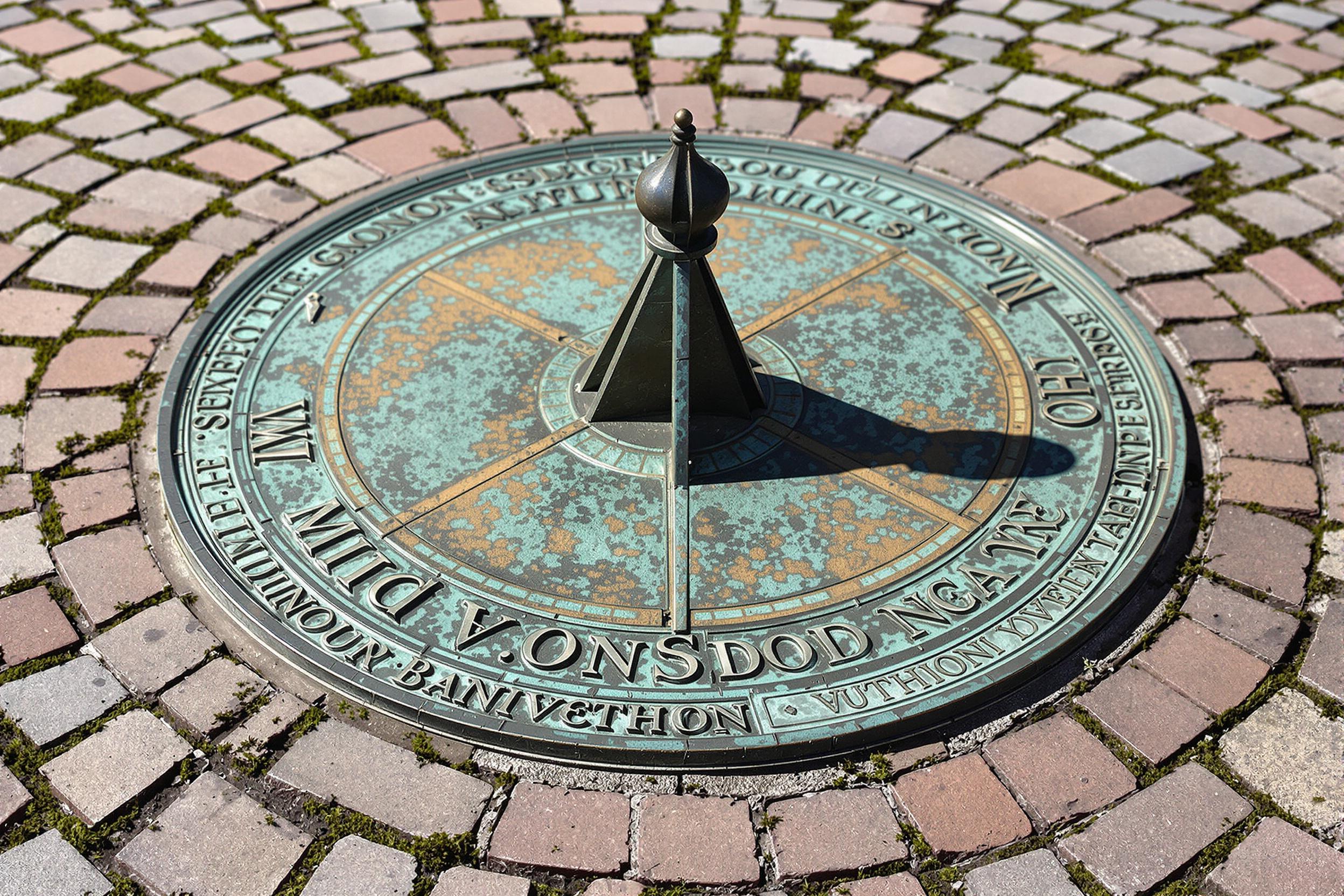 A detailed image of a weathered bronze sundial with intricate carvings atop uneven cobblestones. The shadow of the gnomon (pointer) points to mid-afternoon, cast under brisk sunlight. Verdigris accents highlight the dial's age, while faint moss traces spread across the bricks.