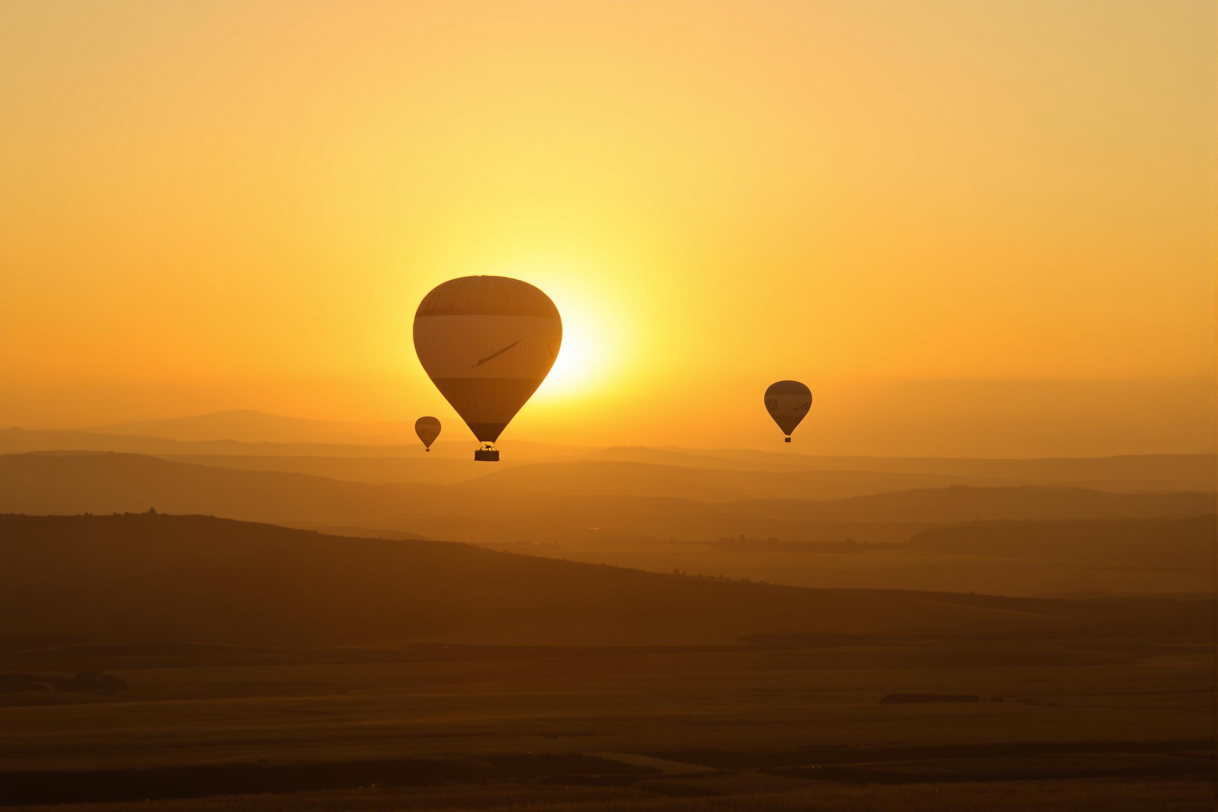 Hot air balloons rise gracefully against a radiant early morning sky. Their dark silhouettes stand in contrast to the warm amber and orange hues of the sunrise. The open field below shows faint details of rolling hills, while the gradient sky transitions from glowing gold to pale blue. A calm and majestic atmosphere dominates the scene.