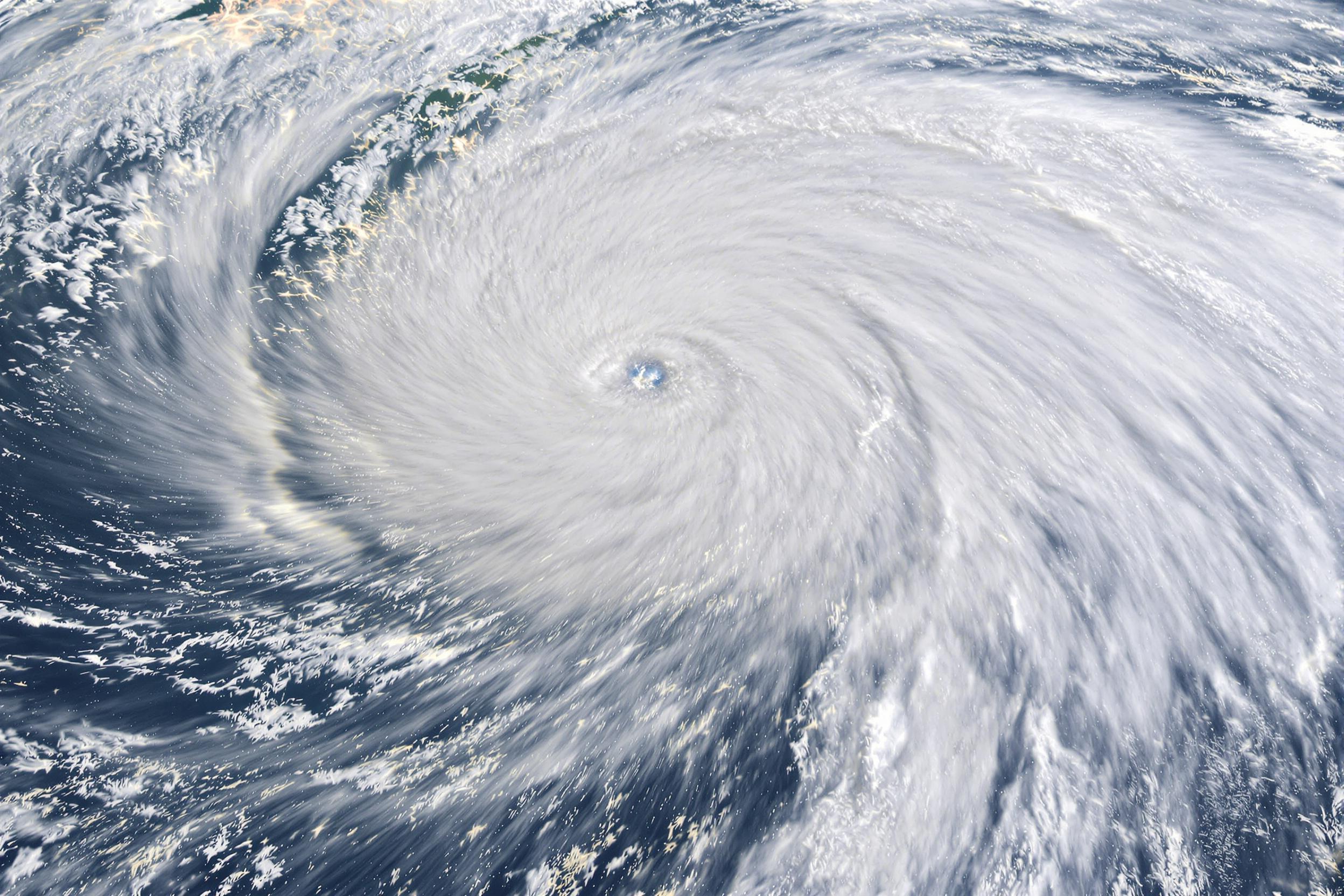 A satellite captures the dramatic spiral of a massive hurricane over azure ocean waters. Crisp, white cloud bands stretch outward, creating mesmerizing swirls around a defined storm eye. Lightning flickers illuminate patches amidst rolling textures. The scene reveals dynamic textural contrasts and vast atmospheric scale under muted blue-green hues.