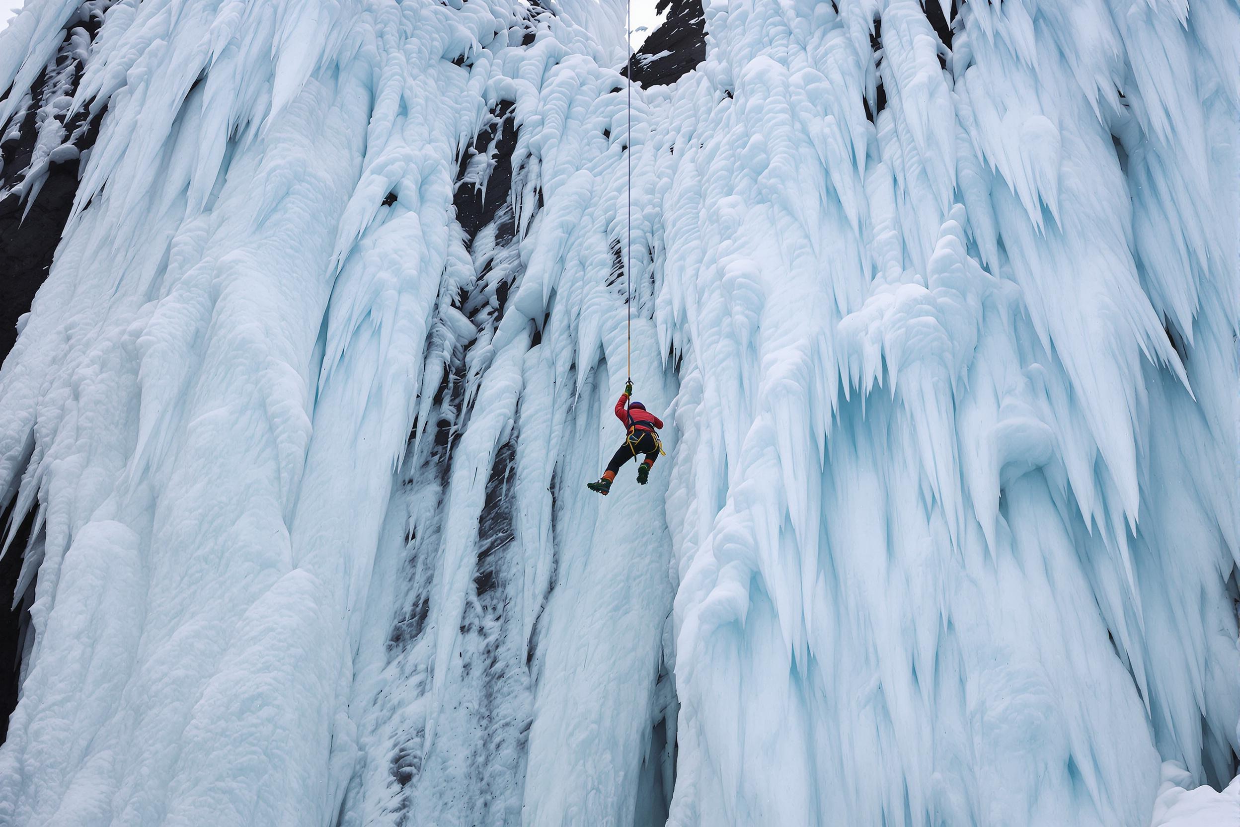 Amid towering icy cliffs, an ice climber hangs mid-ascent, suspended by ropes and spiked boots. The frozen cascade glistens with shades of blue and white as diffused cloudy light captures its intricate details. The climber’s brightly colored jacket contrasts sharply against the monochromatic, wintry backdrop, creating a dynamic focal point.