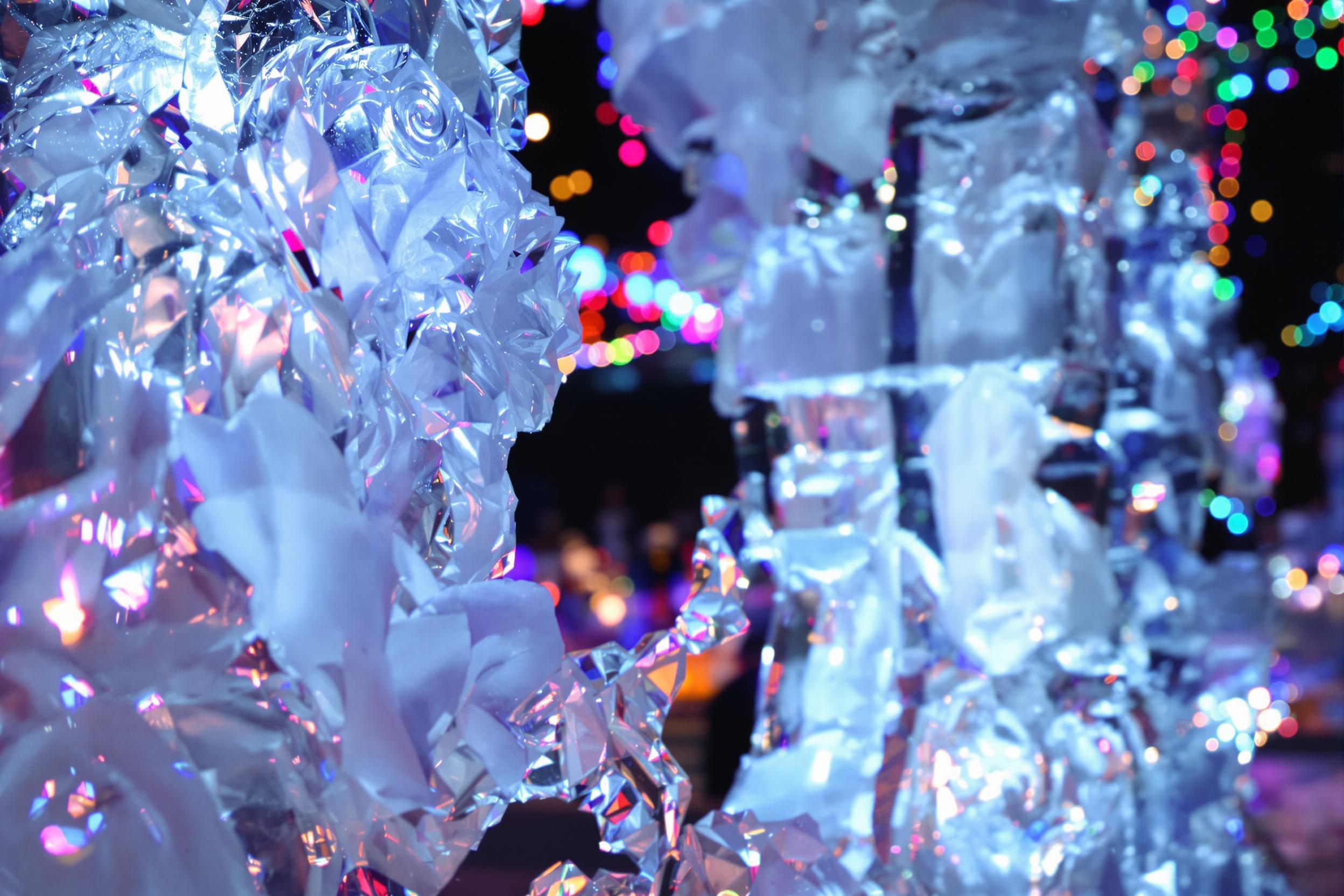 A close-up captures an elaborate ice sculpture illuminated by colorful festival lights. Intricate carvings mimic delicate flowers and swirling patterns, glistening as they catch the light. The surrounding blurred atmosphere hints at a bustling winter festival, enhancing the enchantment of this frosty artwork.