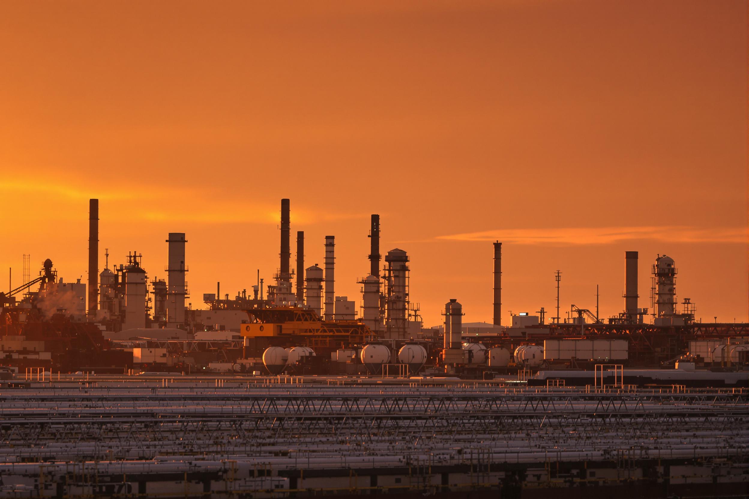 In the golden hues of dusk, an expansive industrial facility stands against a dimming sky colored in radiant warm shades. Smokestacks emit subtle plumes of steam over rows of neat barges and towers. Soft, atmospheric lighting emphasizes the gleaming steel textures of machinery, powered by the day’s receding golden light and incoming depth of evening shadows.