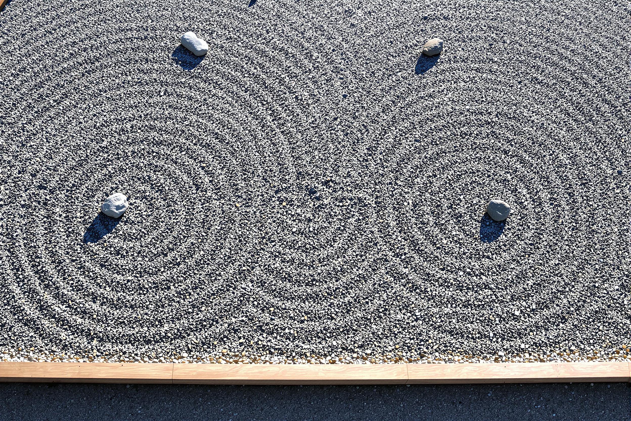 A meticulously arranged Japanese rock garden seen from above showcases symmetrically raked gravel patterns punctuated by carefully placed stones. Sunlight highlights the contours in the raked designs, creating defined shadows. A simple wooden border frames the tranquil scene, emphasizing balance and harmony.