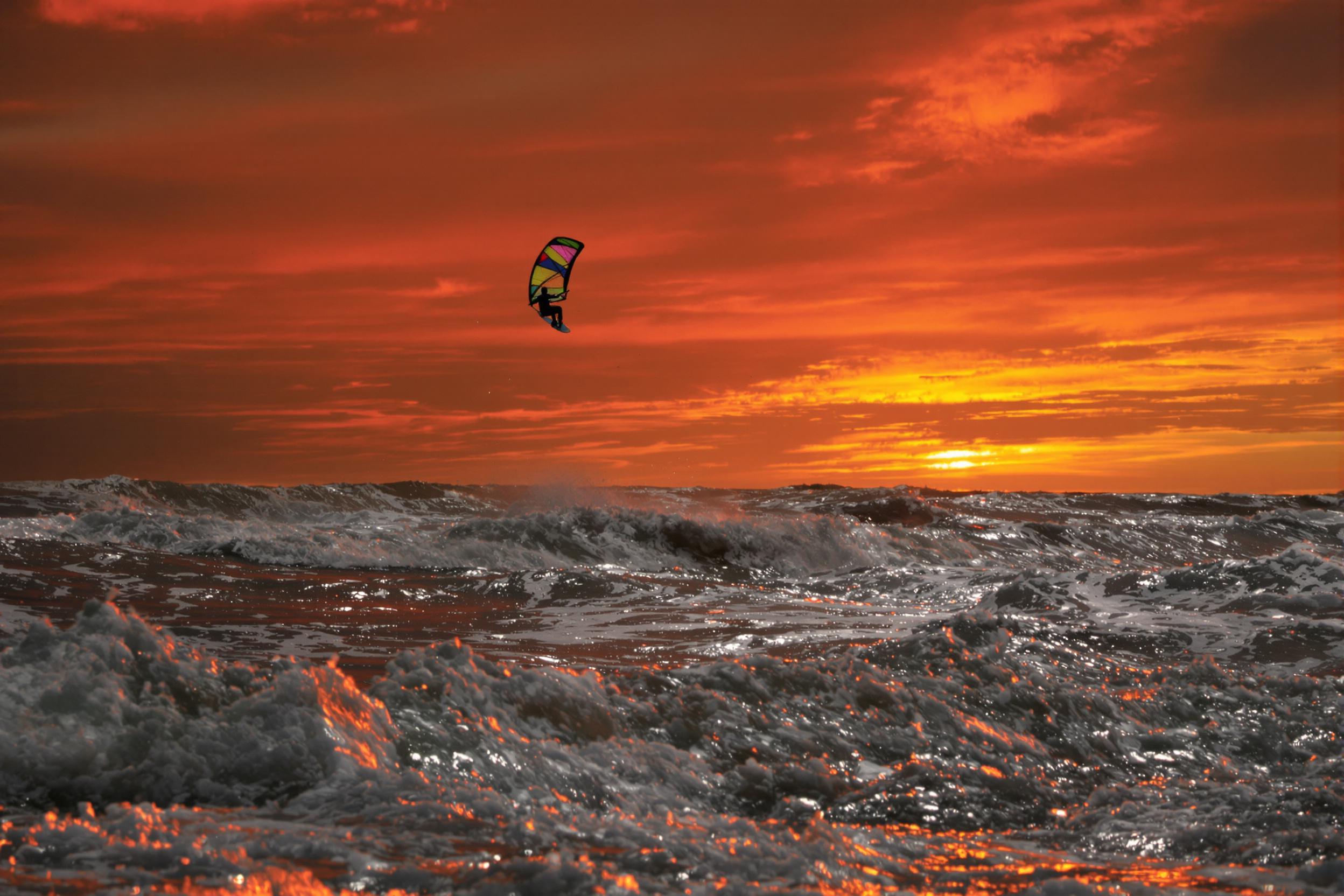 Against a vibrant coastal sunset, a kiteboarder soars above frothy waves, the orange sky reflecting on glittering water. The taut sail contrasts against the horizon, capturing energy mid-motion. Golden-pink light highlights the athlete’s silhouette, blending the thrill of action with serene natural beauty.