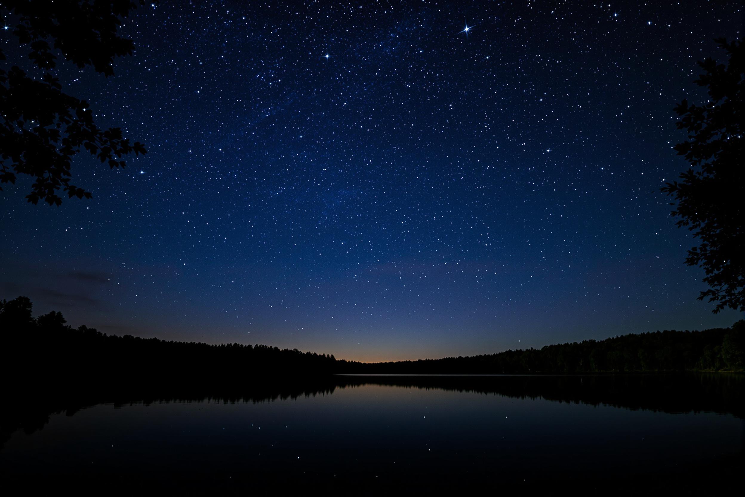 A serene lakeside scene unfolds under a starry night sky. Bright constellations twinkle against deep indigo hues, mirrored perfectly on the lake's glassy surface. Silhouettes of surrounding trees frame the composition, adding to the peaceful ambiance. The stillness is palpable, inviting quiet reflection and admiration for the beauty of the cosmos.