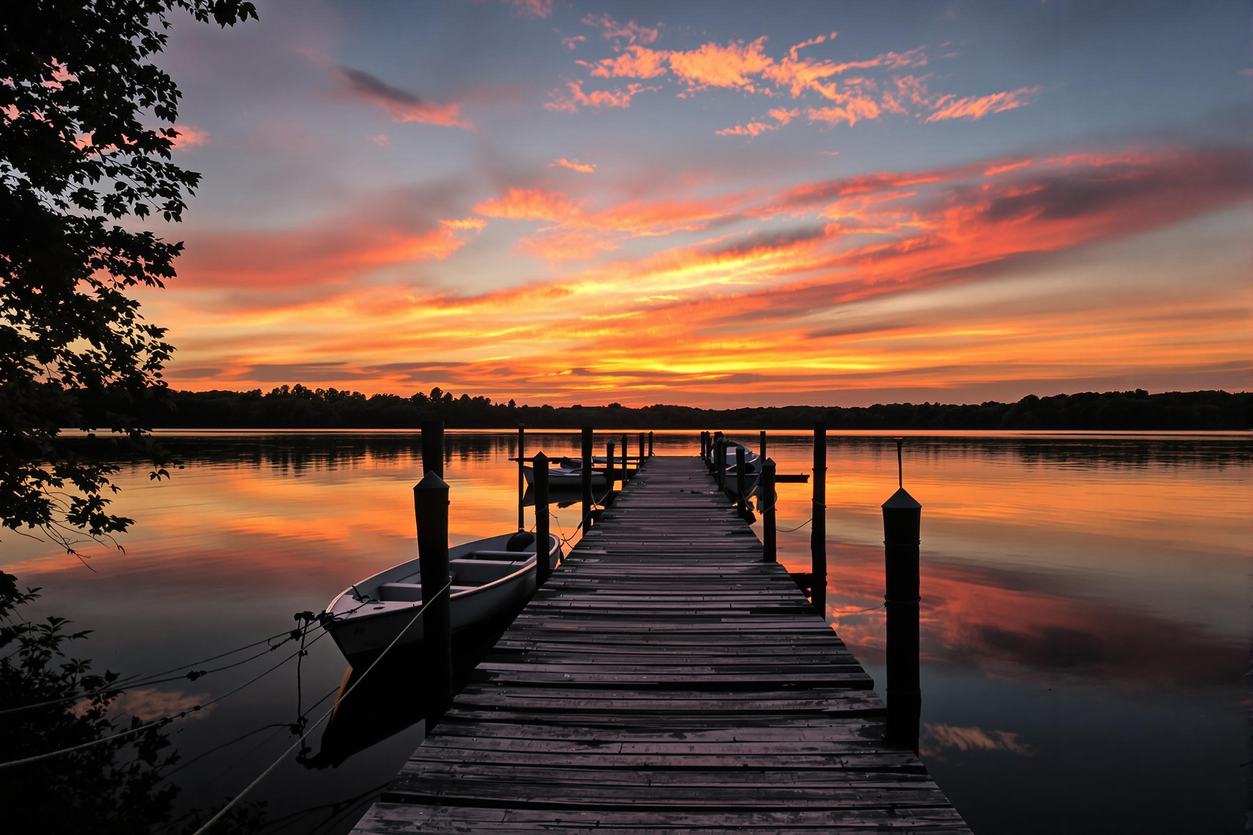 A tranquil lake scene displays multiple boats tethered to a weathered wooden dock as the sun sets. Warm oranges and pinks blend beautifully across the horizon, reflected in the smooth water's surface. Silhouettes of distant trees frame the scene, while wisps of clouds catch the vibrant colors above, enhancing the idyllic setting.