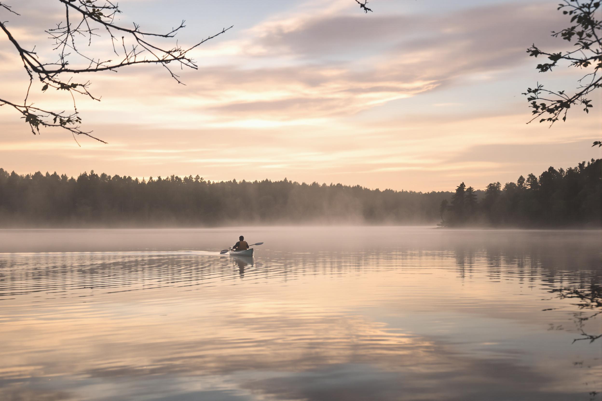 A tranquil lake emerges under the soft glow of early dawn. A solitary canoeist glides silently through the misty waters, creating gentle ripples that distort the reflected palette of pastel-colored skies. Surrounding trees stand still, their silhouettes framed against the fading darkness as the first light of day slowly illuminates the serene landscape.