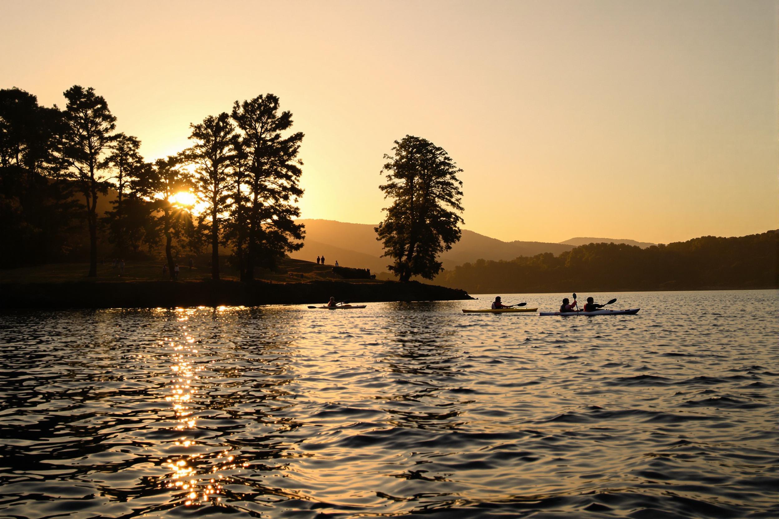 A serene lakeside unfolds at sunset as golden light stretches across the water. Silhouettes of kayakers glide gently through rippling waves, their paddles breaking the glassy surface. Towering trees frame the shore, their leaves kissed by the evening glow. Hills rise in the background, softly blending into hues of orange and pink, creating a tranquil atmosphere.