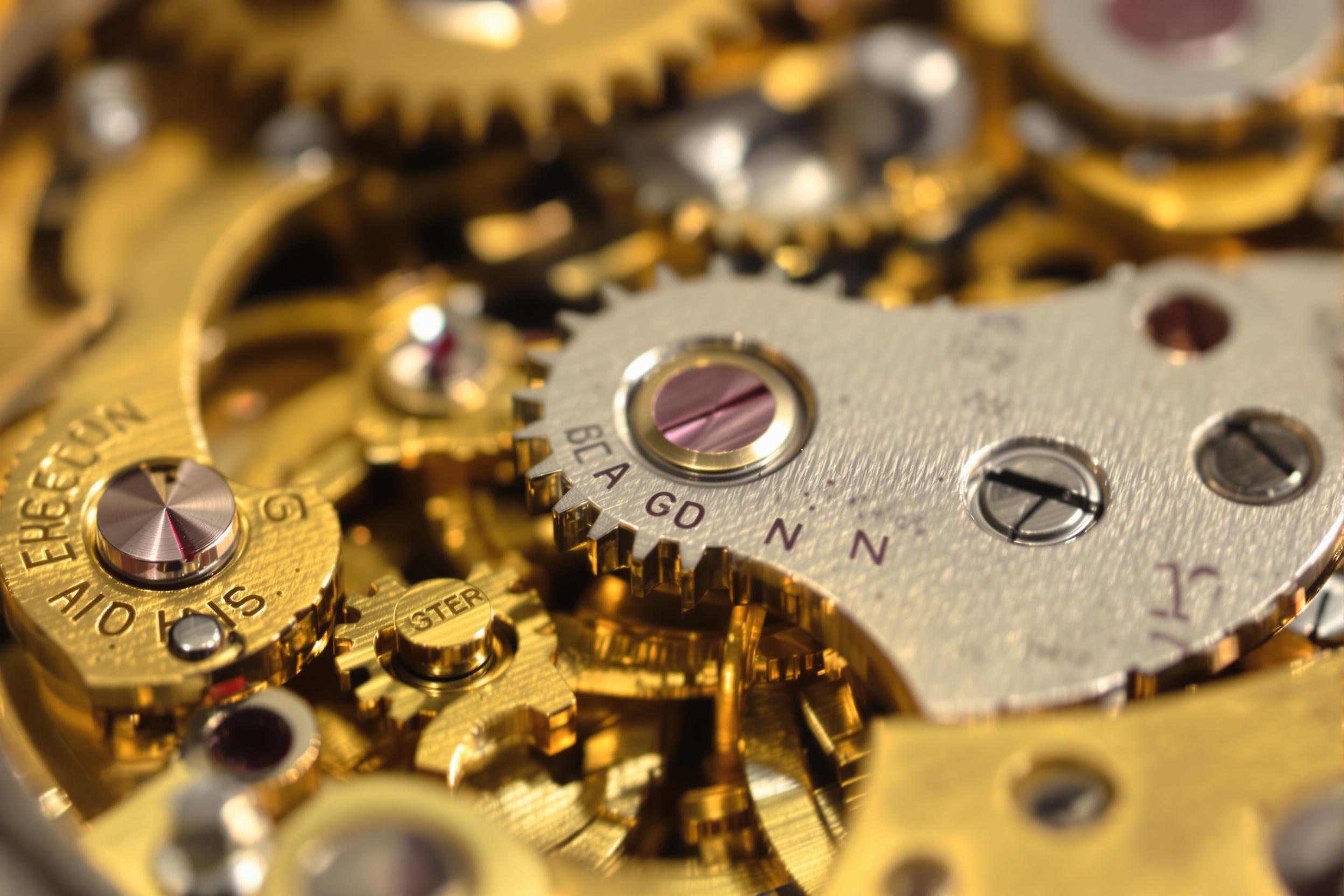 A macro photograph focuses on the intricate mechanics of a vintage pocket watch. Polished metallic gears and springs interlock precisely, their golden and silver hues glowing under diffuse, warm lighting. Fine scratches and patina give character to the aged metal. Soft shadows and reflective accents enhance depth in this captivating composition.