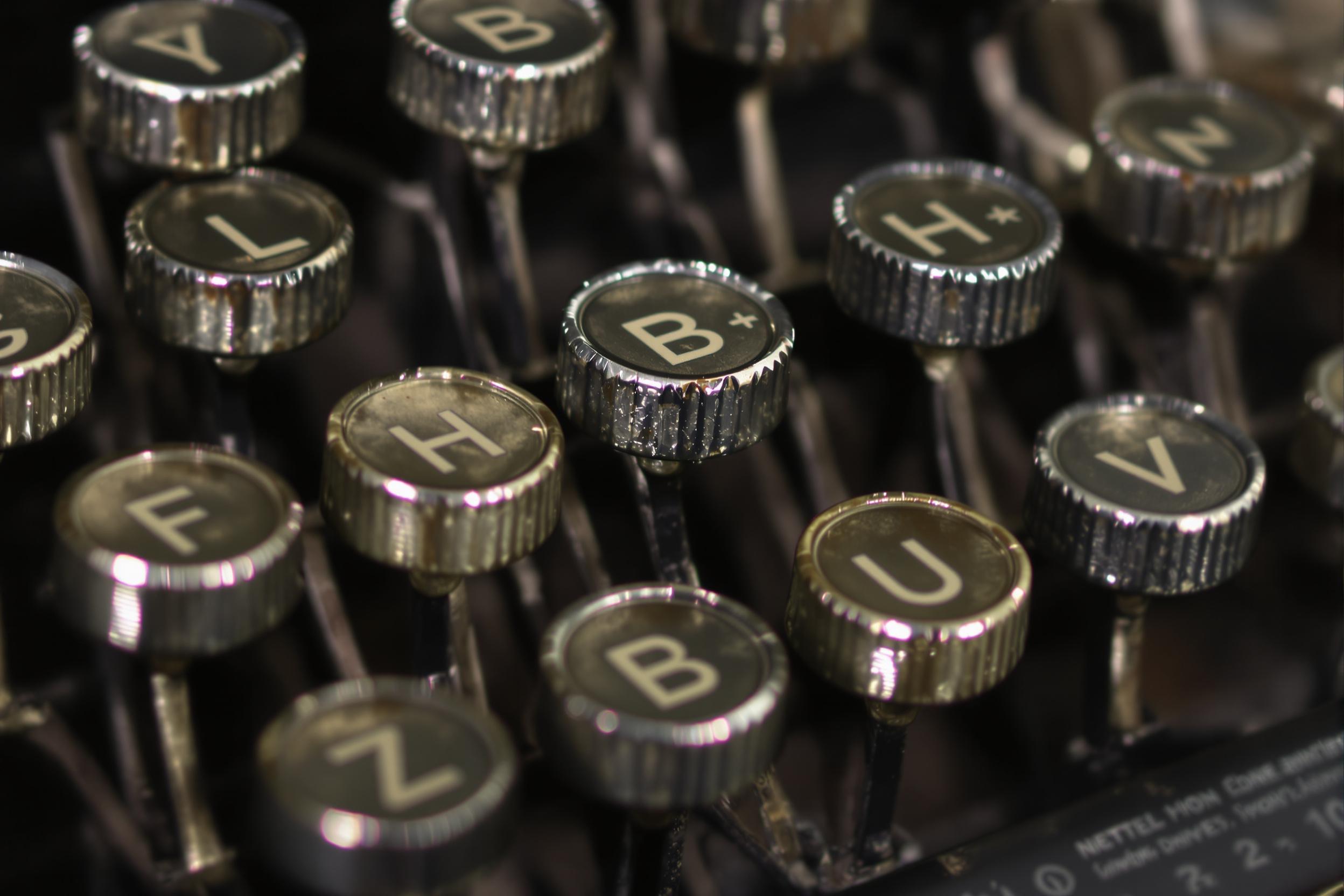 An ultra-close macro image showcases the individual metal typebars of a vintage typewriter. Each letter key, worn from use, reveals intricate grease patterns and faint scratches that catch diffused ambient light. The muted silver sheen of the metal contrasts against matte black surfaces in shallow focus, accentuating textural nuances.