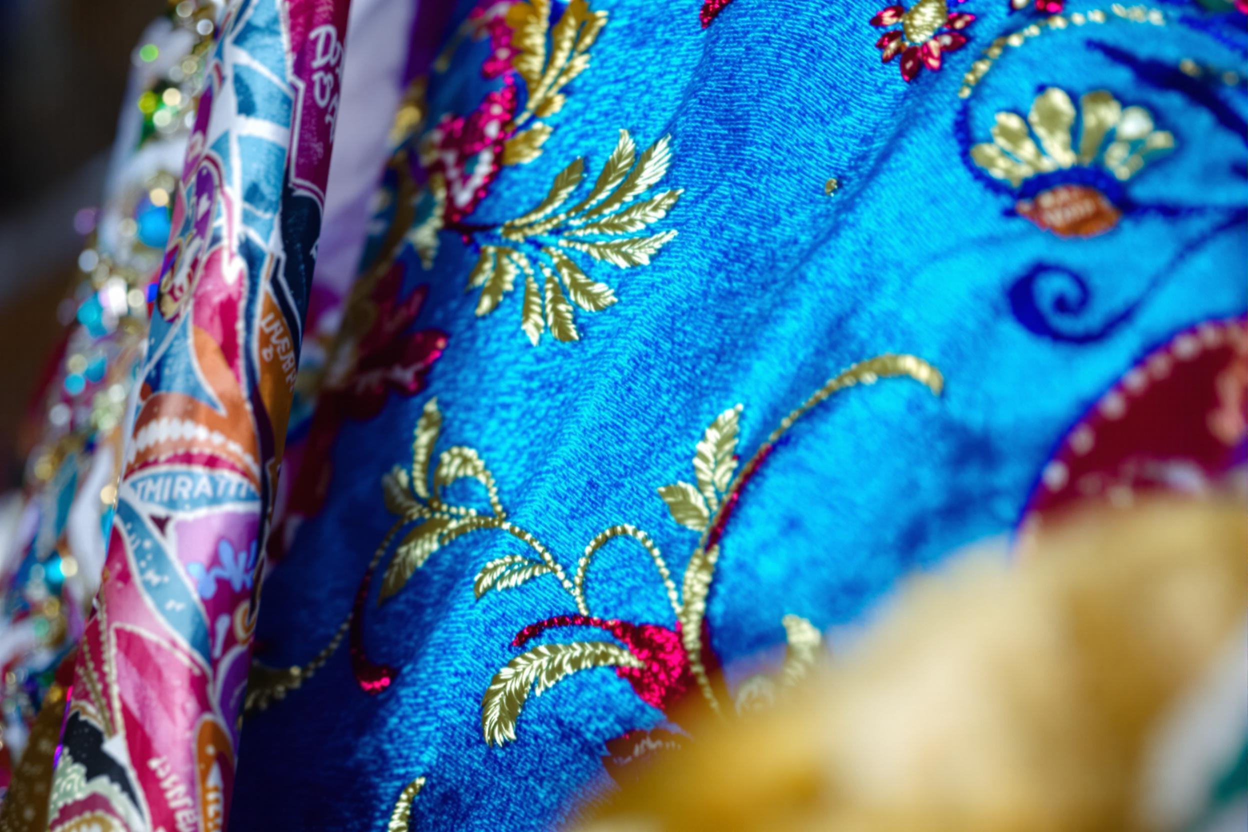 A close-up reveals the intricate beauty of traditional hand embroidery on a vivid fabric. Delicate threads in vibrant shades of blue, red, and gold create a floral motif. The stitched surface is illuminated softly, enhancing the texture against blurred edges framed by background textiles.