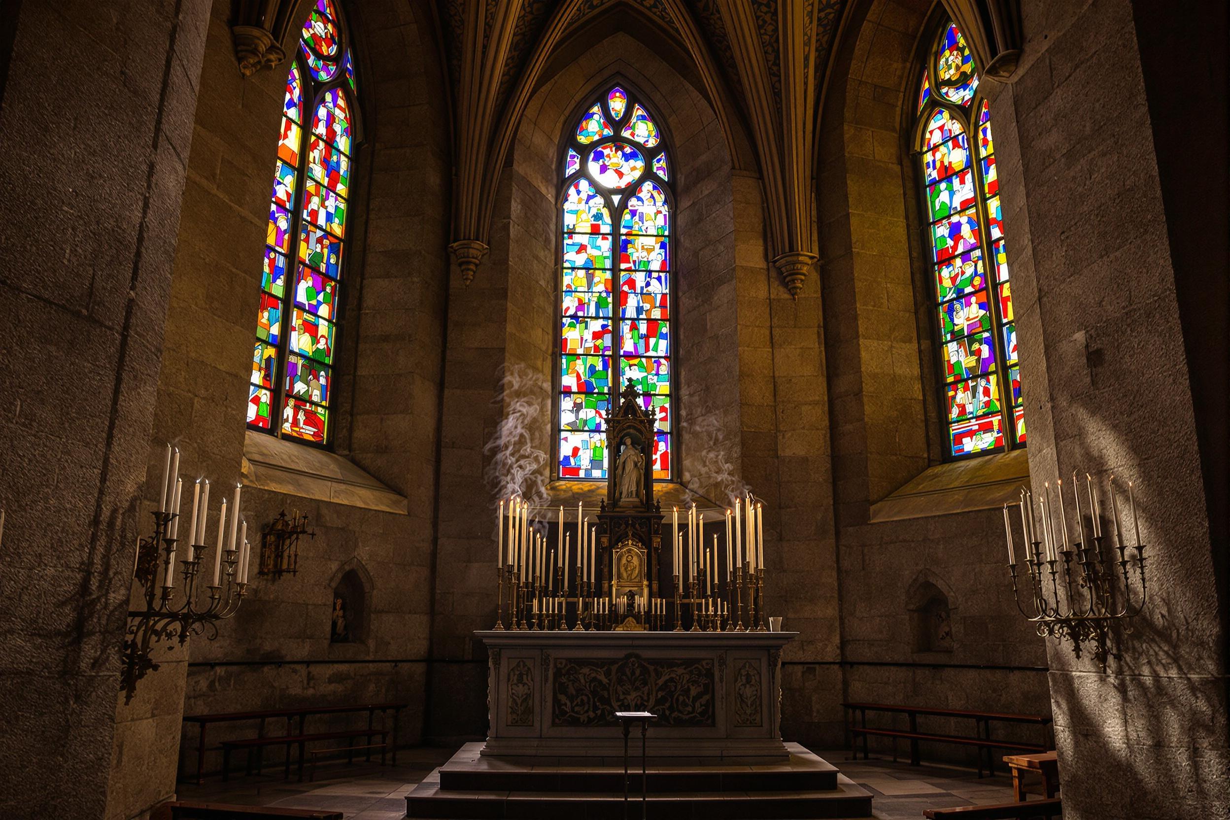 Inside a medieval stone cathedral, slender candles line a carved altar under multi-hued stained glass windows. Incense rises in soft wisps, filling the dimly lit air with motion. Colored sunlight filters through the ornate glass, casting vivid patterns across ancient textured walls and worn flagstones, showcasing timeless serenity.