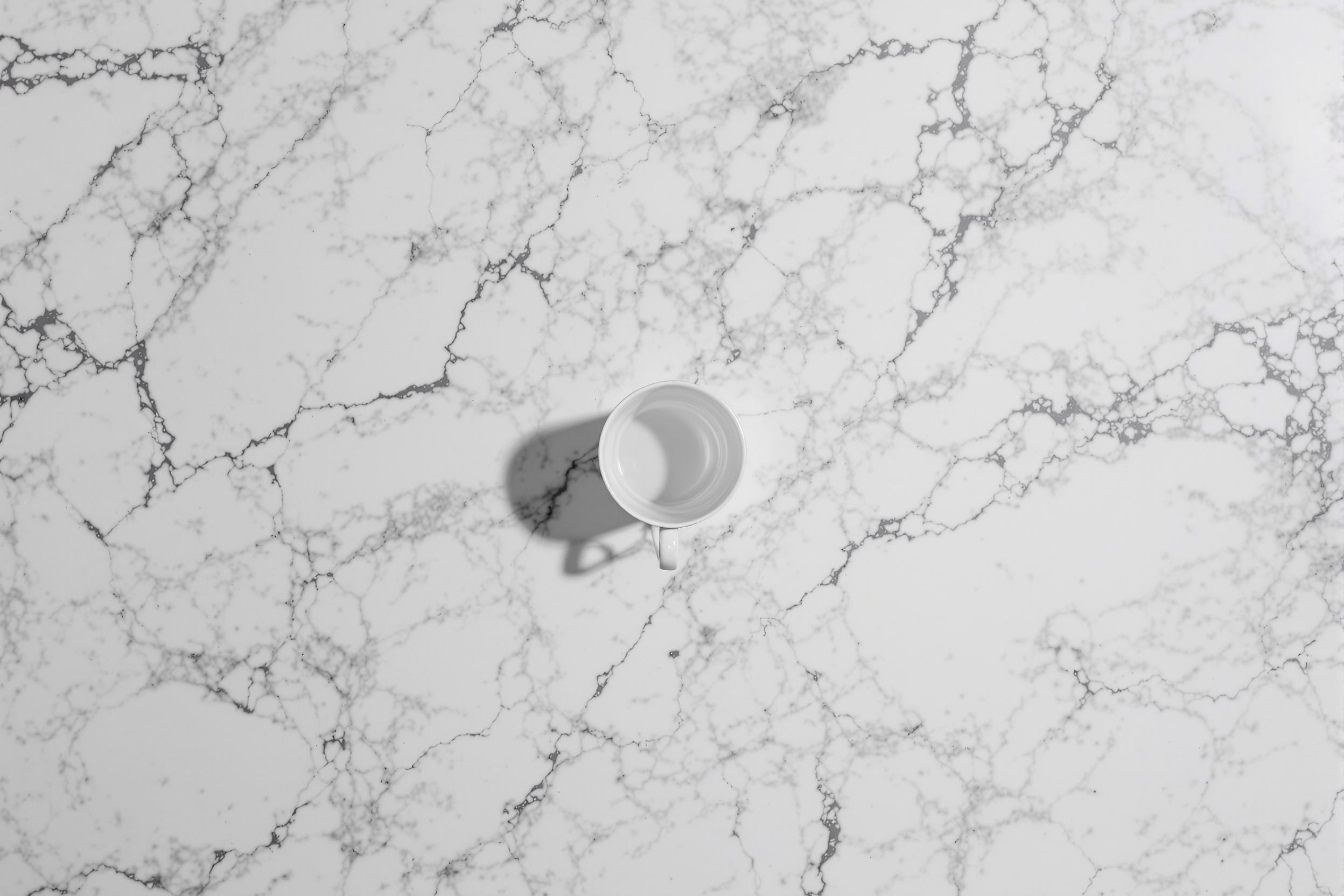 A single white porcelain teacup sits centered on a vast expanse of veined marble. The cup's delicate handle casts a subtle shadow, creating a study in simplicity and contrast. This minimalist composition showcases the beauty of negative space and refined elegance.