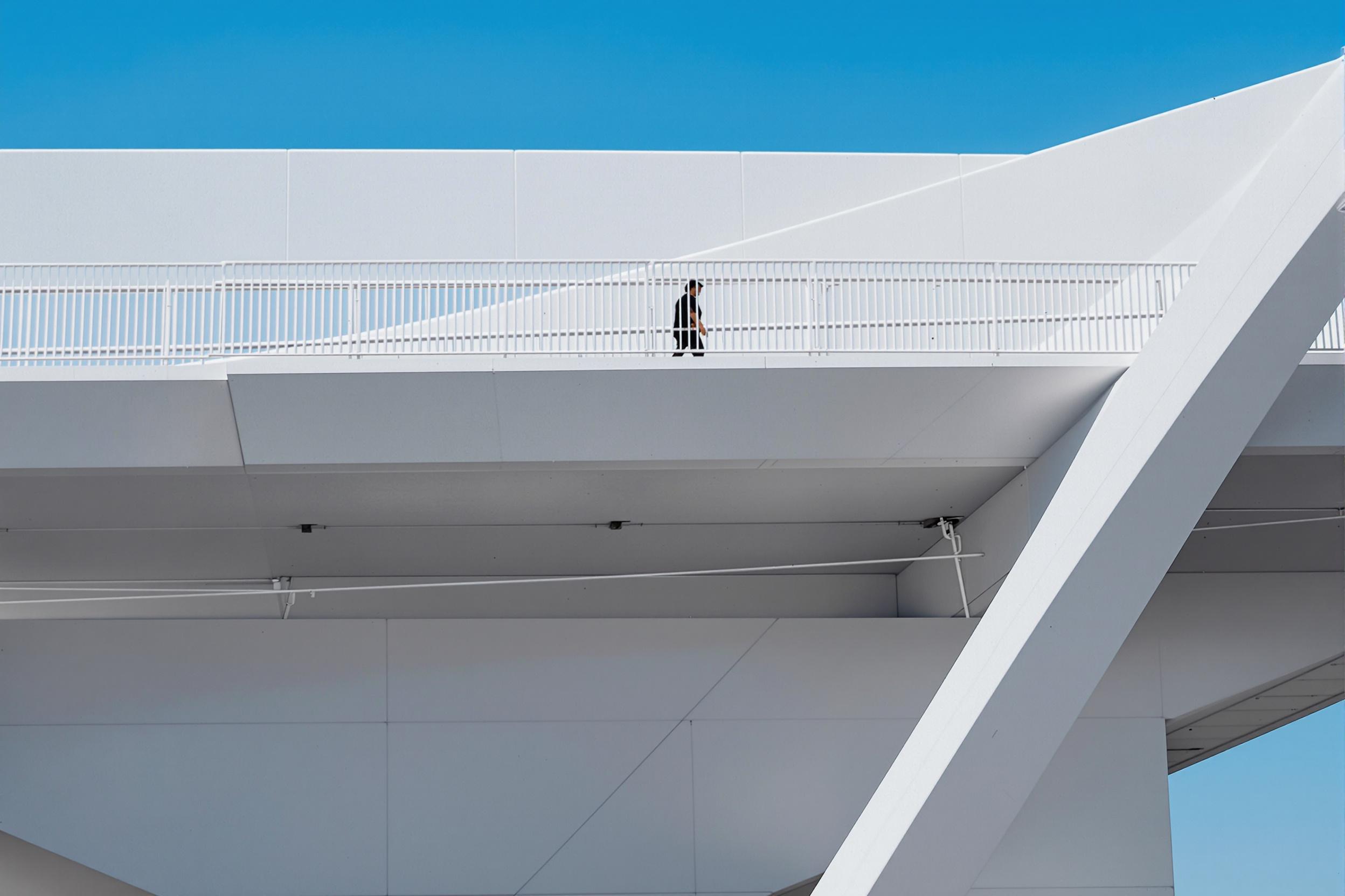 Lone figure walking across a stark white pedestrian bridge against a clear blue sky. Clean lines and geometric shapes create a powerful minimalist urban landscape.