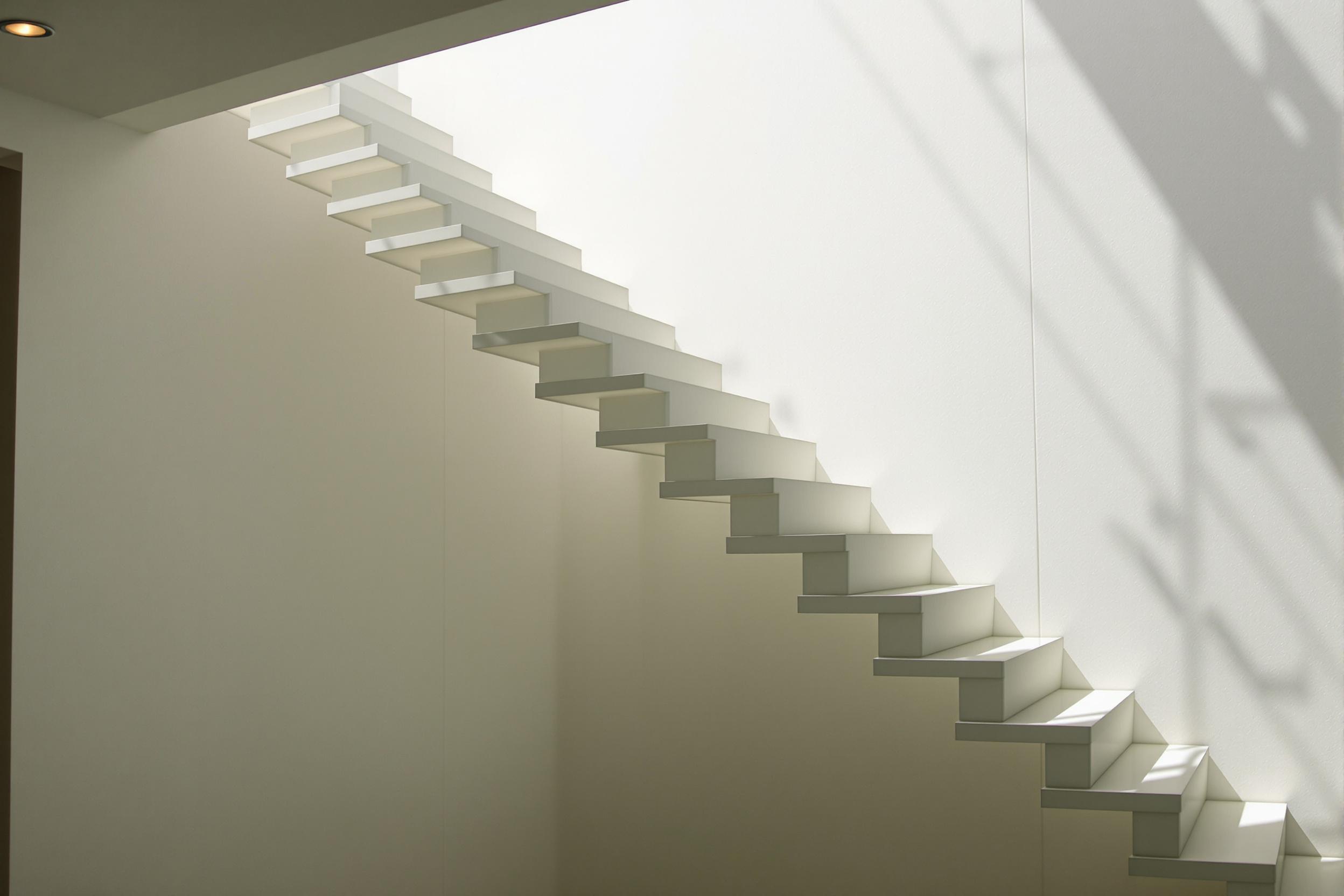 A sleek, minimalist floating staircase ascends through a sun-drenched atrium. White treads appear to hover, casting crisp shadows on the bare wall. Natural light streams in, creating a play of light and shadow in this serene architectural space.