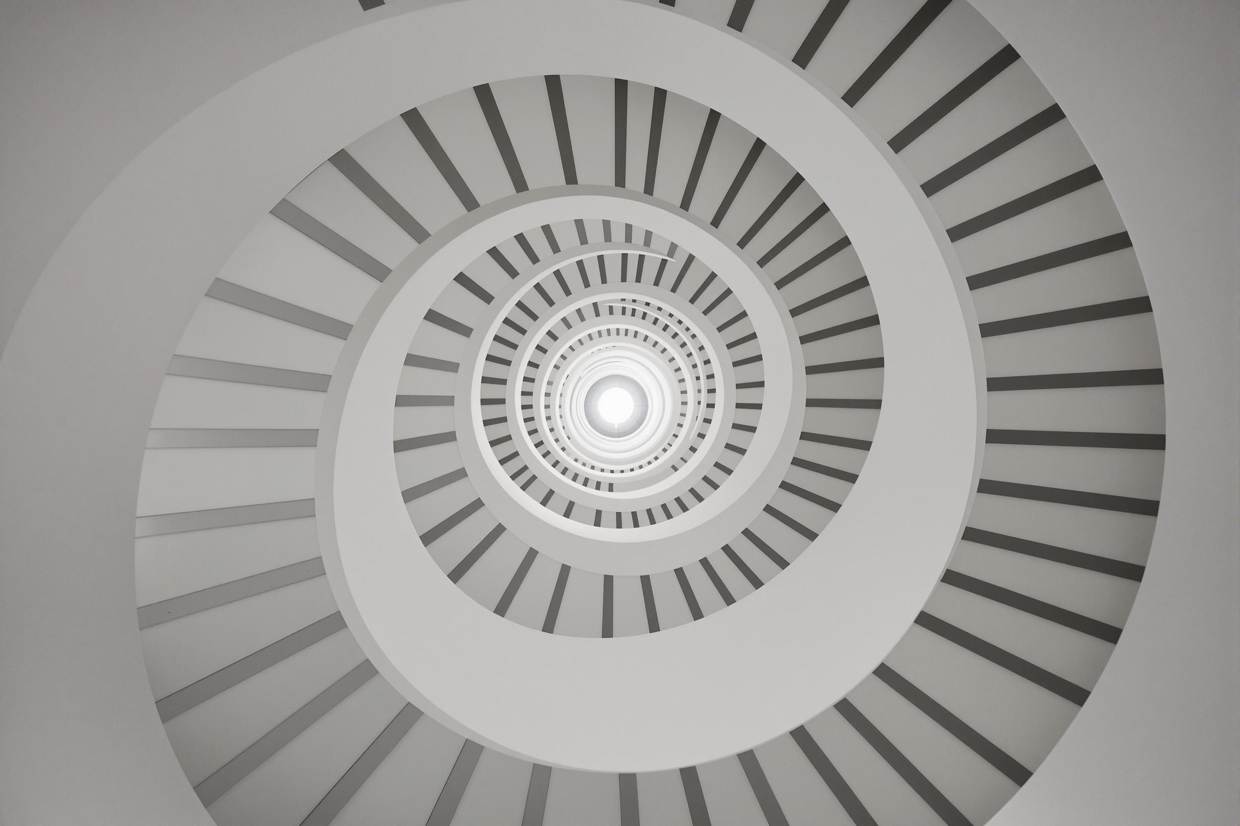 A striking minimalist staircase spirals upward, its clean lines and monochromatic palette creating a bold architectural statement. The structure's geometric precision and flowing curves showcase the beauty of simplicity in modern design.