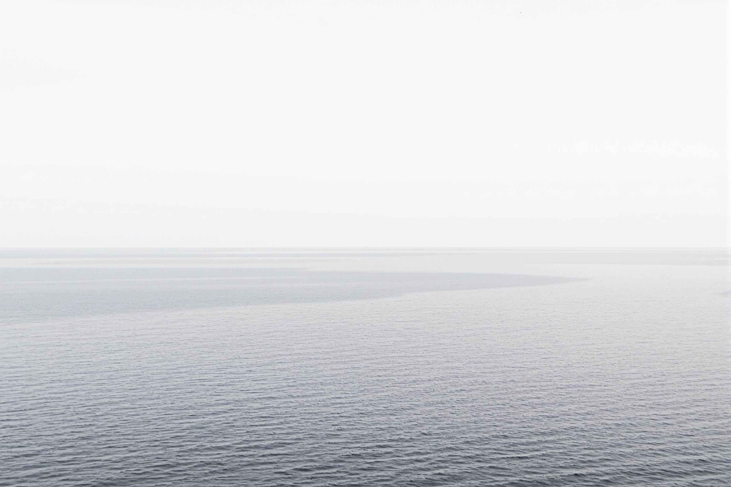 A serene minimalist seascape where the ocean seamlessly blends with the sky at the horizon. The composition features a vast expanse of calm water in muted blue-gray tones, occupying the lower two-thirds of the frame. The upper third showcases a cloudless sky in a slightly lighter hue, creating a subtle gradient effect. The horizon line is perfectly straight and centered, dividing the image into two distinct yet harmonious sections. This minimalist photograph captures the essence of tranquility and infinite space, inviting viewers to contemplate the beauty of simplicity in nature.