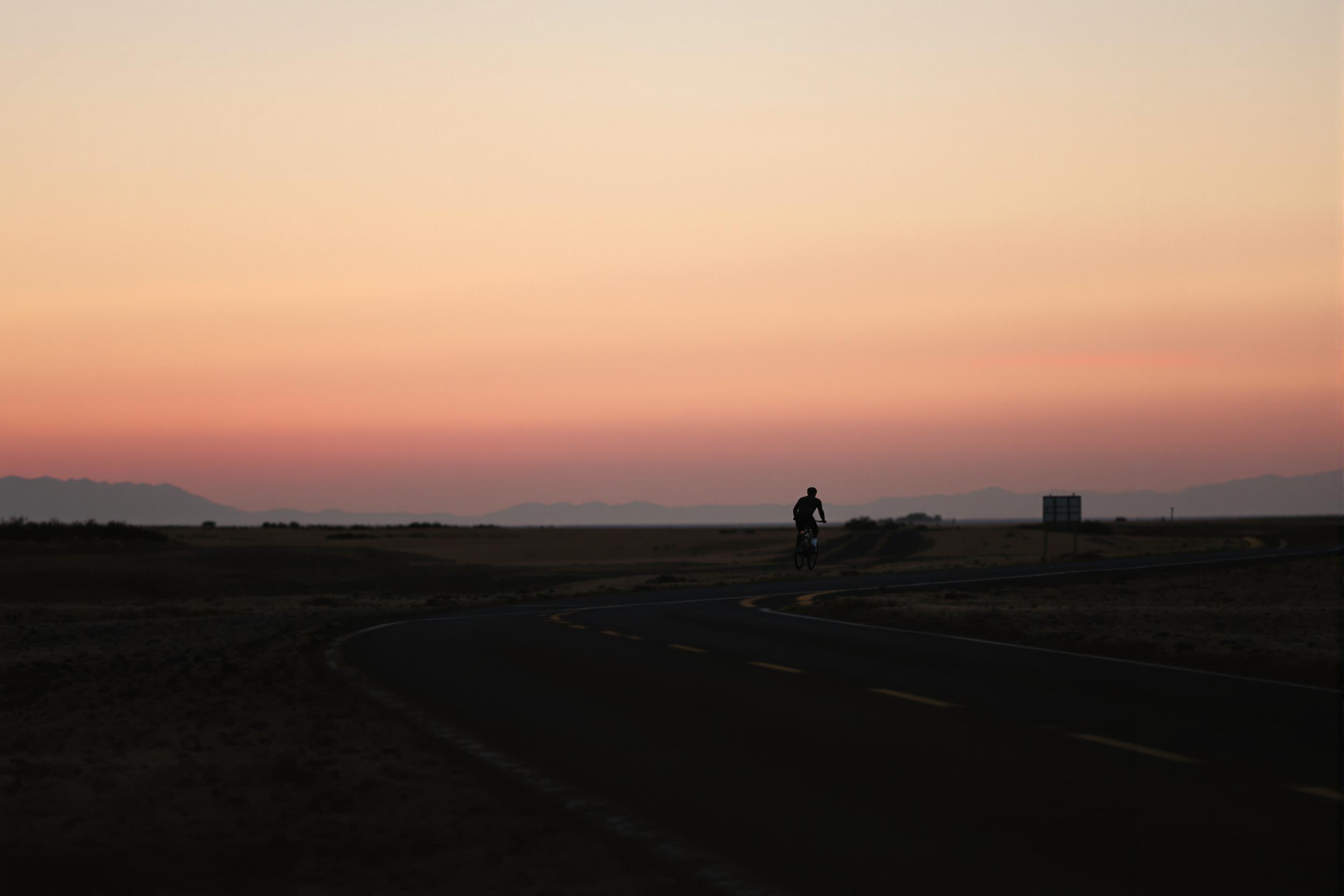 A solitary cyclist pedals along a deserted road, silhouetted against a pastel sky at dawn. The image captures the essence of solitude, freedom, and the open road in a striking minimalist composition.