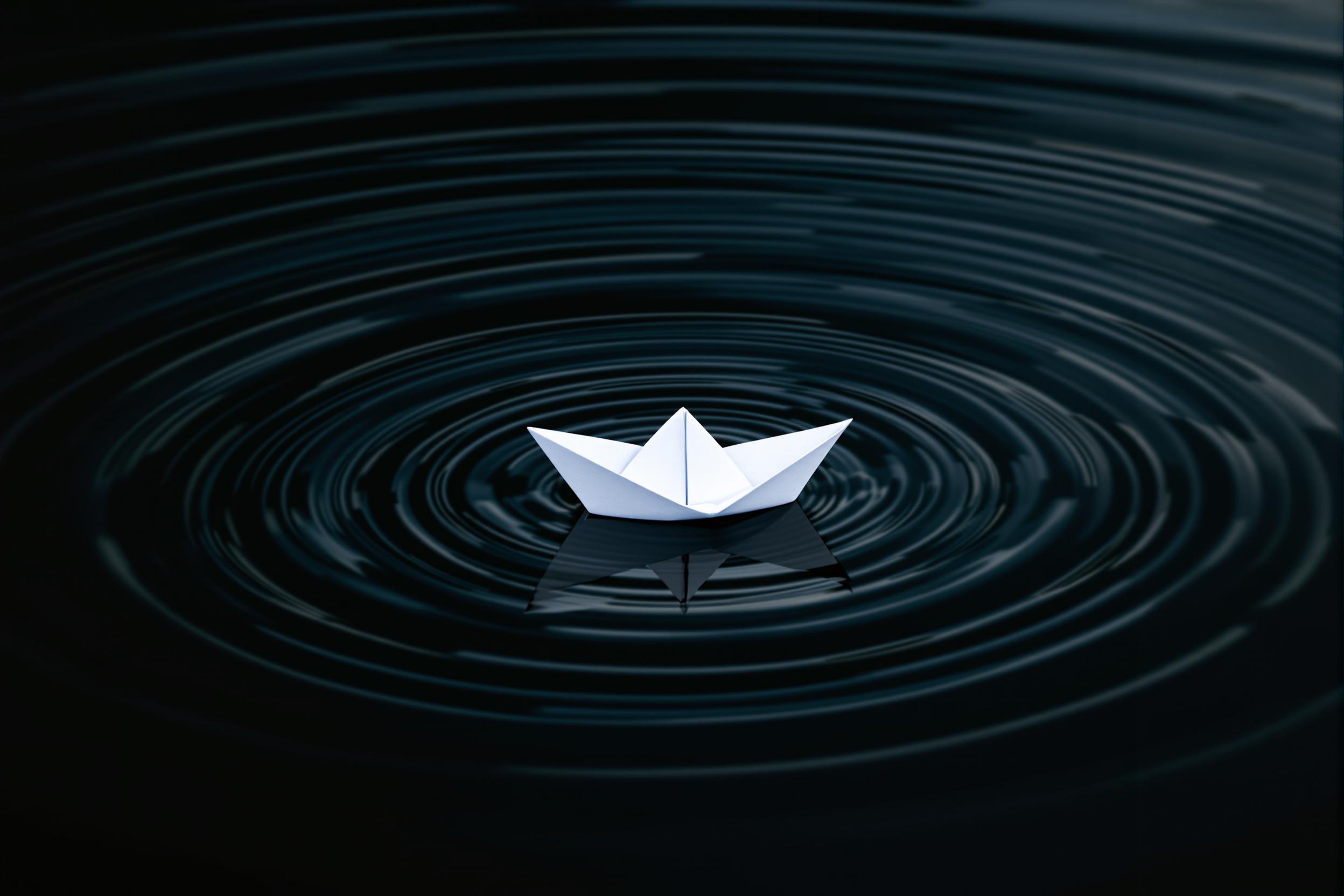 A delicate white paper origami boat floats on gently rippling water, creating concentric circles. The image captures the interplay of simplicity and movement, showcasing minimalist design principles in a serene aquatic setting.