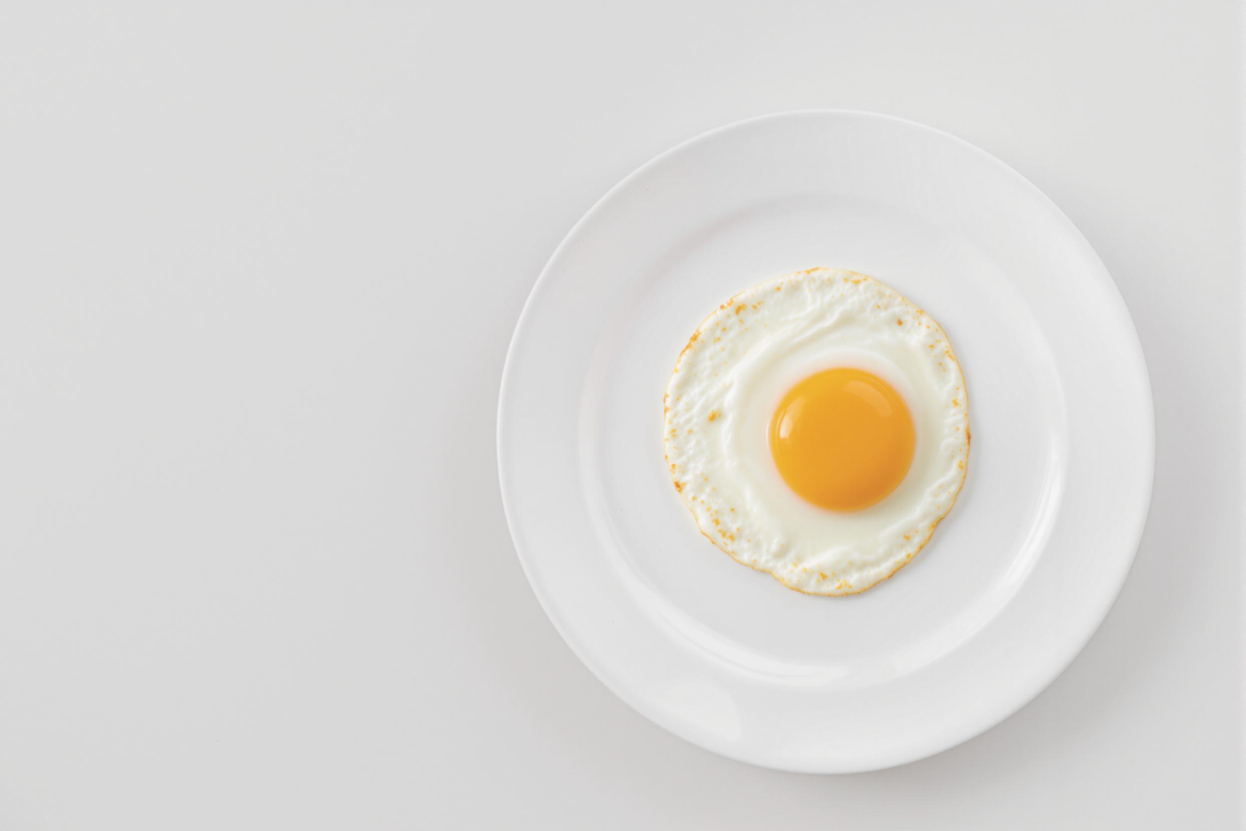 A solitary perfectly fried egg rests on a pristine white plate, set against a light gray background. The yolk's vibrant yellow creates a striking contrast, embodying simplicity and culinary elegance in this minimalist food photography composition.