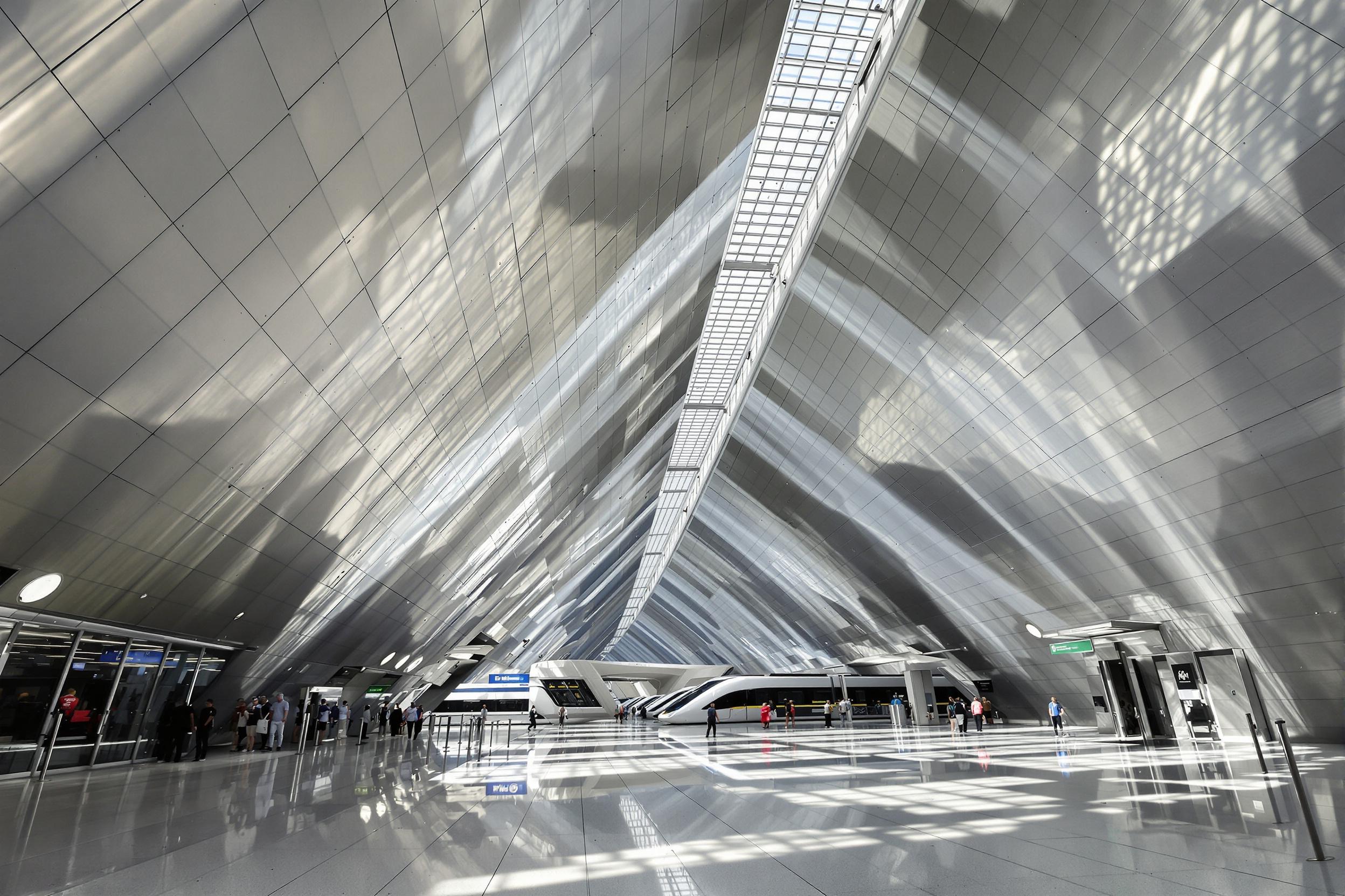 A sleek, modern train station showcases bold futuristic architecture under bright daylight. Narrow beams of sunlight stream through intricate grid-like skylights onto polished floors below. Metallic, curved walls and sweeping diagonals dominate the composition, while small figures traverse the vast space, adding scale.