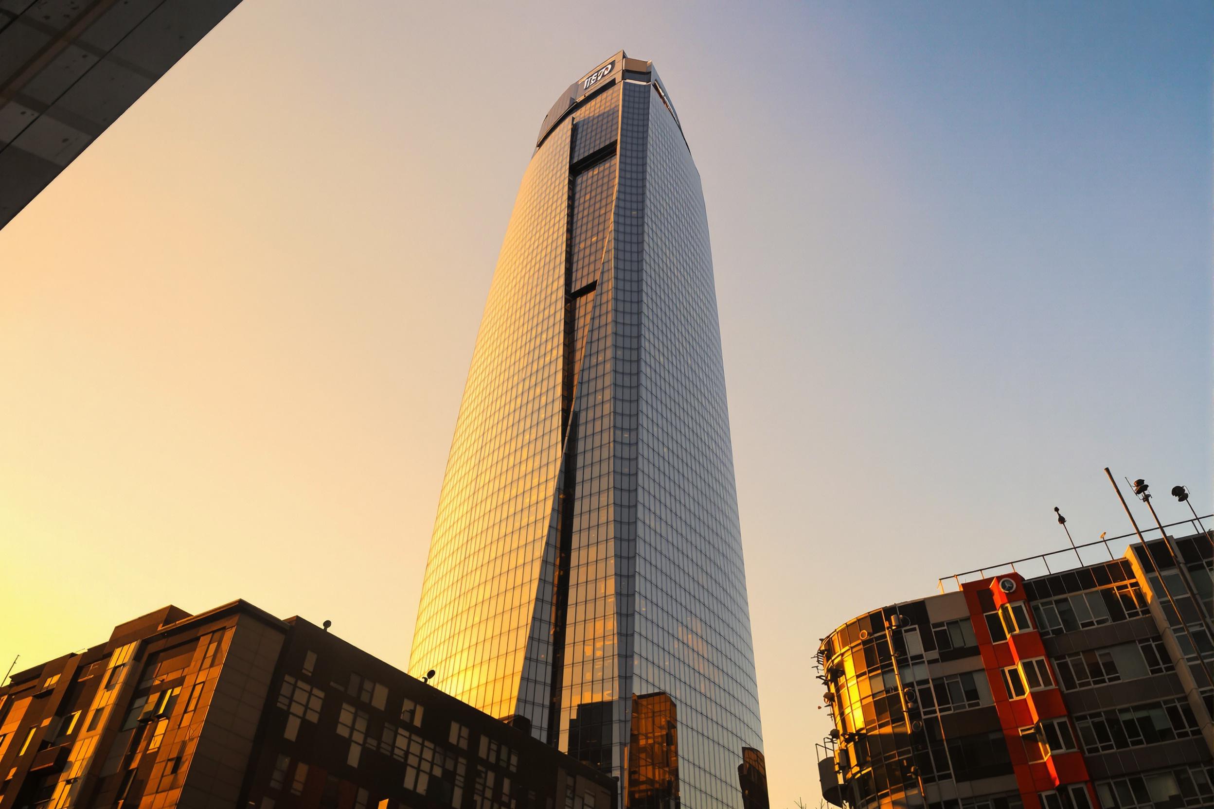 As the sun rises over a modern city, its golden light reflects vividly across a glass skyscraper's towering facade. The sleek lines of the building stretch upward against a pale orange and blue gradient sky. Shadows from neighboring architecture frame the scene, enhancing the interplay of light and urban textures.