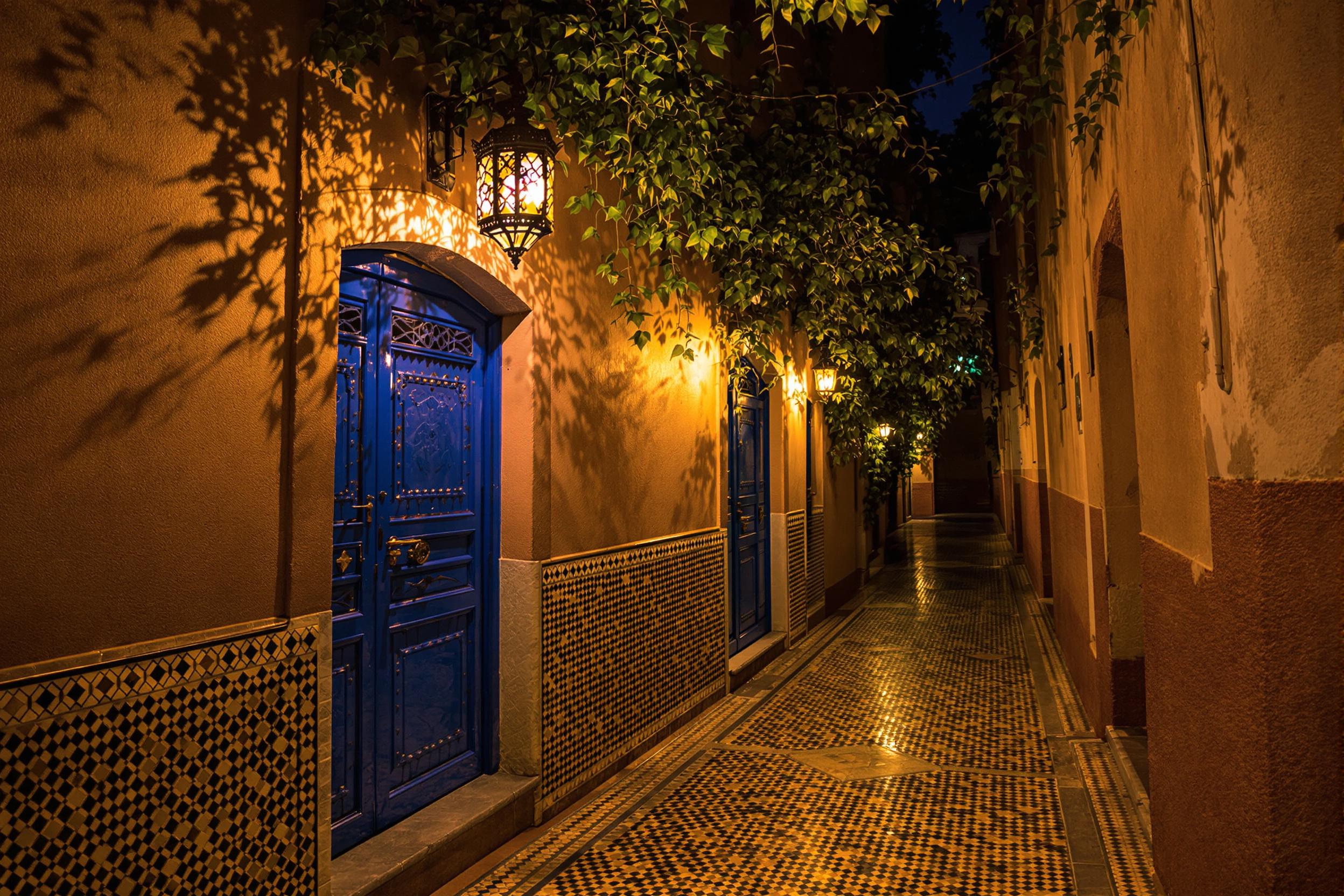 A narrow Moroccan alley glows under artfully carved lantern light reflecting warm amber hues onto decorative mosaic tiles. The aged stucco walls display vibrant cobalt blue doors with ornate ironworks. Golden-toned shadows softly fill this dreamy nighttime passage framed by hints of lush climbing ivy overhead.