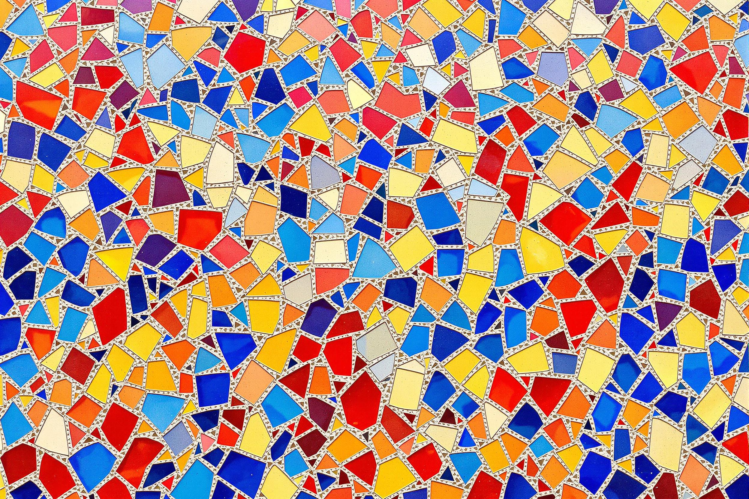 A close-up study of an intricate full-wall mosaic made of multicolored tiles. Each tile is irregularly shaped, fitting together seamlessly to form vivid, abstract patterns of fiery reds, cobalt blues, and golden yellows. Subtle grout lines between the tiles add texture under bright, even sunlight, which enhances the vibrancy of the artwork. The overall composition emphasizes balanced geometry and artistic detail.