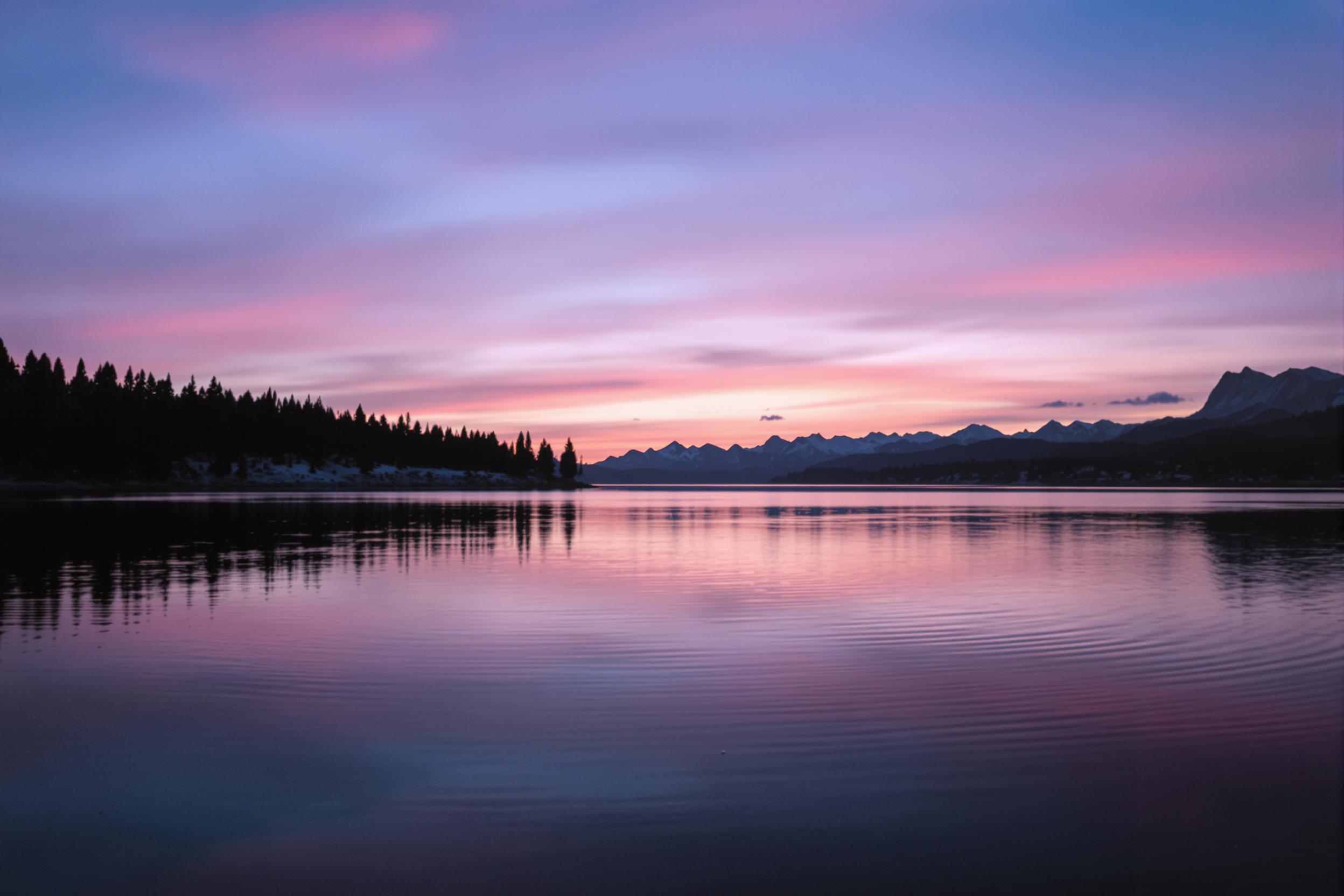 A tranquil mountain lake reflects the soft hues of twilight as the sun sets behind distant peaks. Silhouetted trees line the shore, their dark forms contrasting against the fading pinks and purples of the sky. The glassy water captures these colors perfectly, enhancing the serene atmosphere. Gentle ripples disturb the reflection, inviting peaceful contemplation.