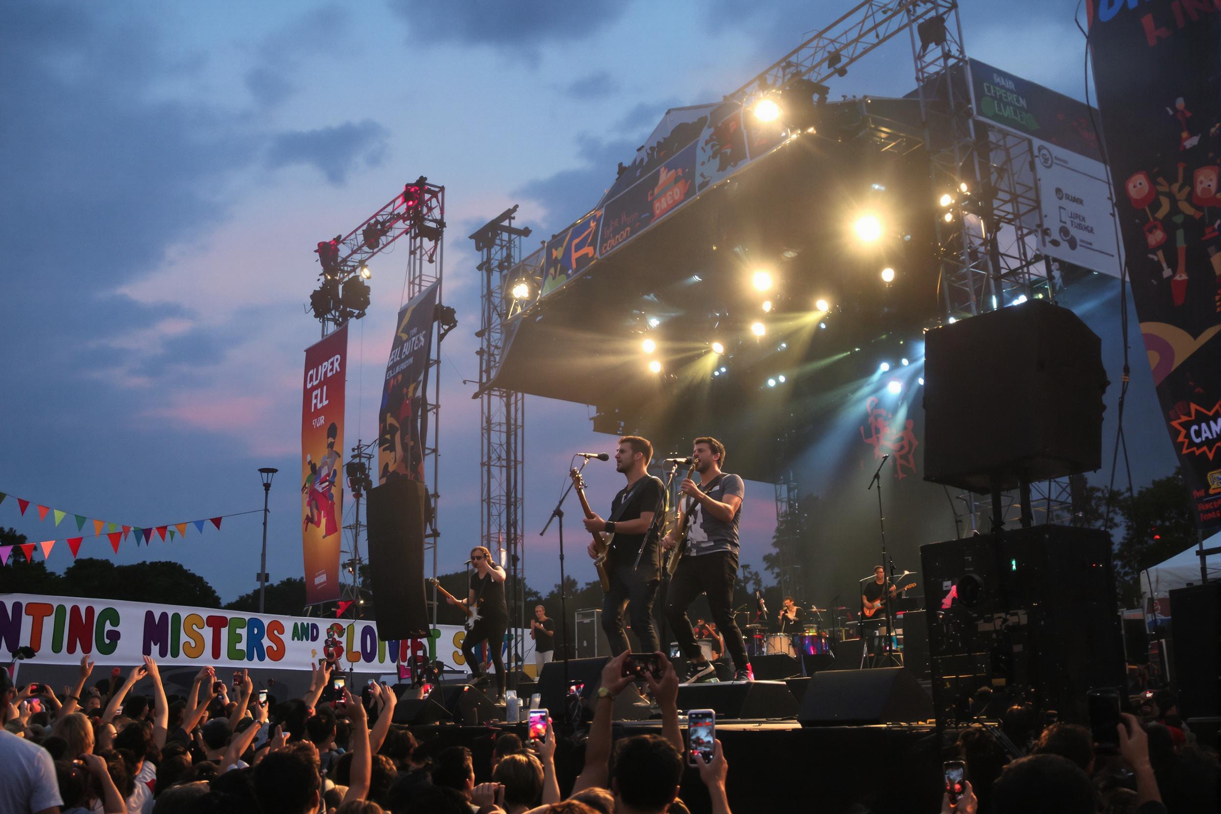 A vibrant outdoor music festival comes alive as a group of musicians performs energetically on stage. The scene captures performers passionately playing various instruments under glowing stage lights against a backdrop of a dusky sky. Excited festival-goers sway with the rhythm, while colorful banners decorate the area.