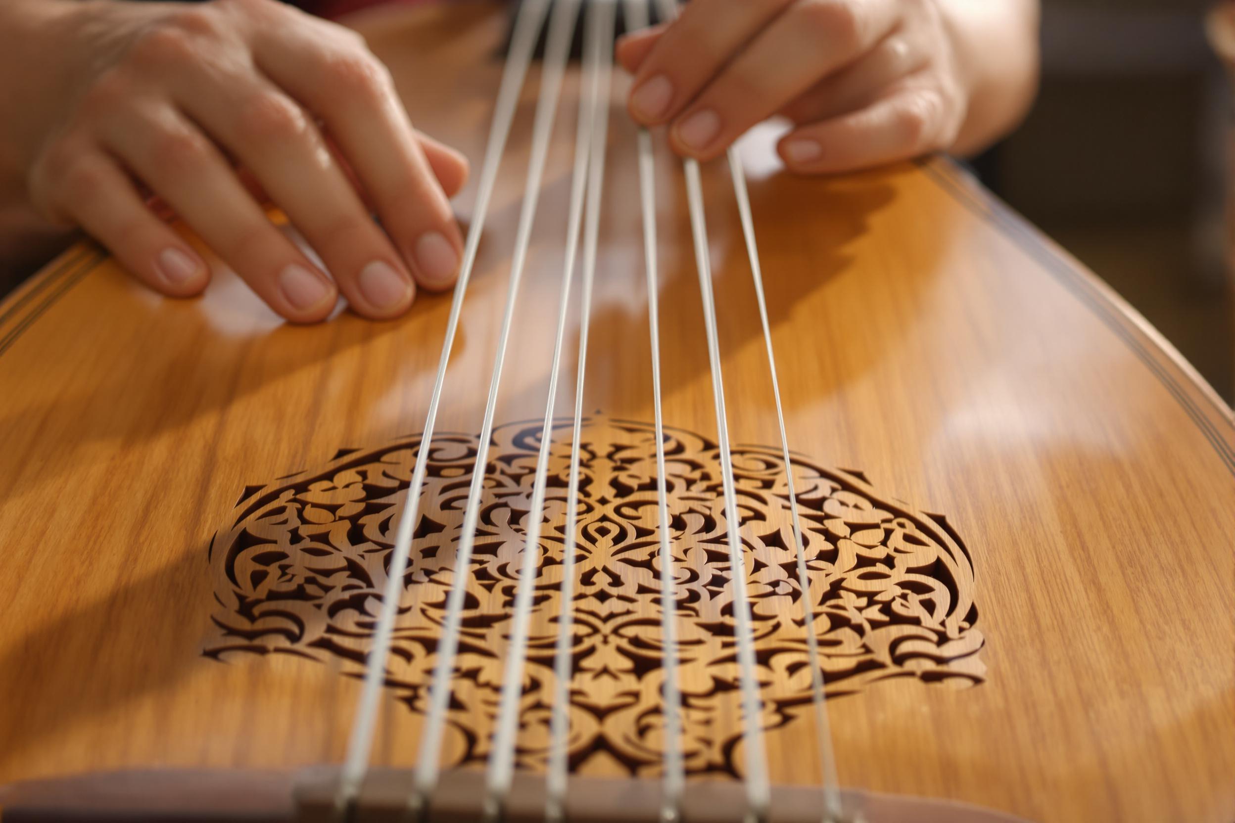 A close-up captures skilled hands elegantly plucking strings on a handcrafted wooden lute inside a softly-lit room. The polished wood gleams with warm, honeyed tones, showcasing delicate carvings on its body. Gentle hues of amber light enhance the texture of the strings and intricate design, evoking a sense of artistry and timeless tradition.