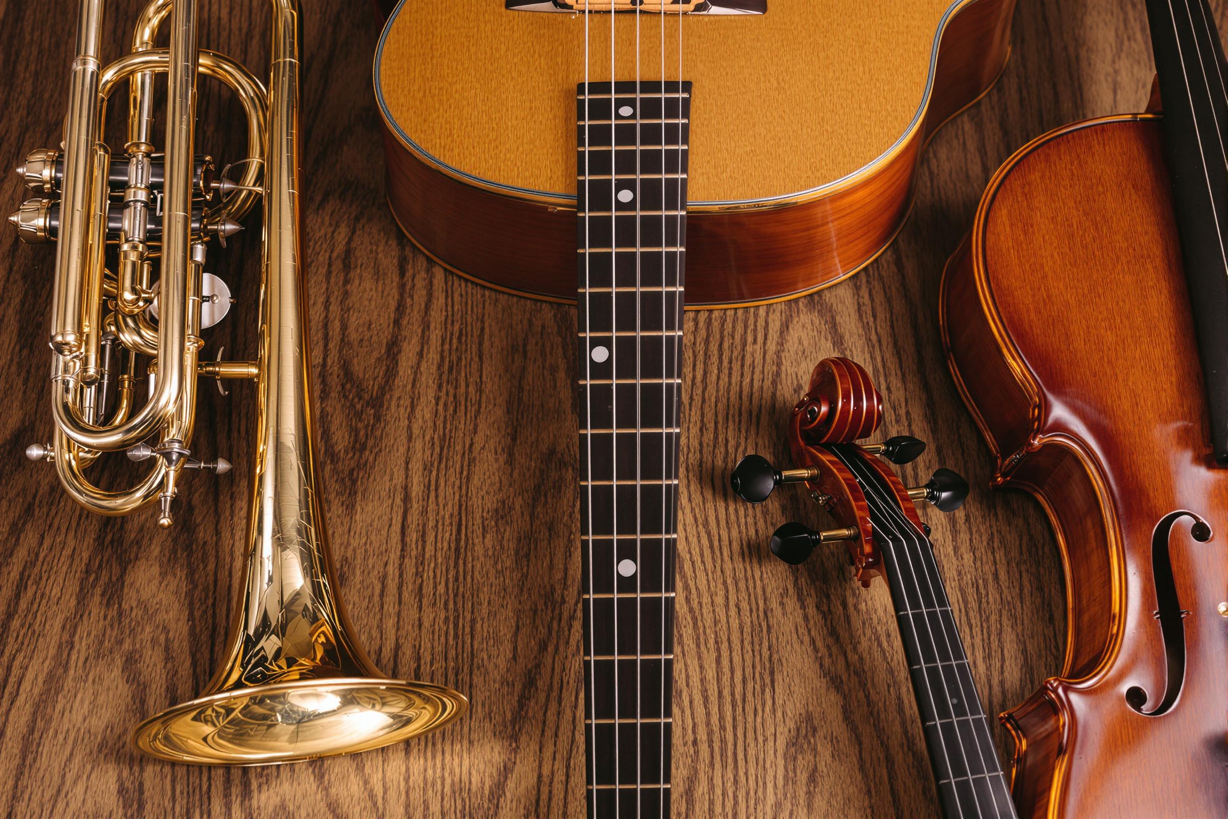 A meticulously arranged close-up captures a collection of diverse musical instruments atop a textured wooden surface. The shiny brass of a trumpet gleams softly alongside the smooth curves of an acoustic guitar, while a pair of elegant violins showcase their polished finishes. A gentle play of light emphasizes the intricate details of each instrument, reflecting a celebration of artistry and craftsmanship.