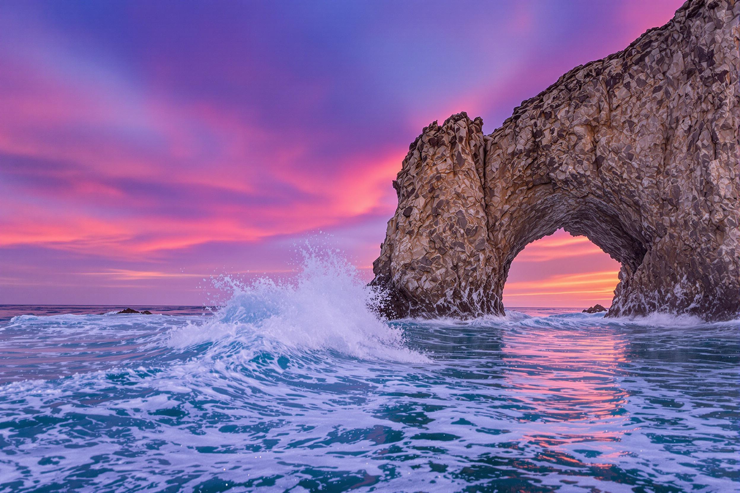 Waves crash dramatically against a massive natural stone arch jutting from crystal-clear sea waters. Vibrant twilight hues—deep lavender, fiery orange, and soft pinks—paint the sky and reflect subtly on rippling surfaces. The unique coastal formation is seen in a wide-framed perspective showcasing its stark contrast against the serene backdrop.